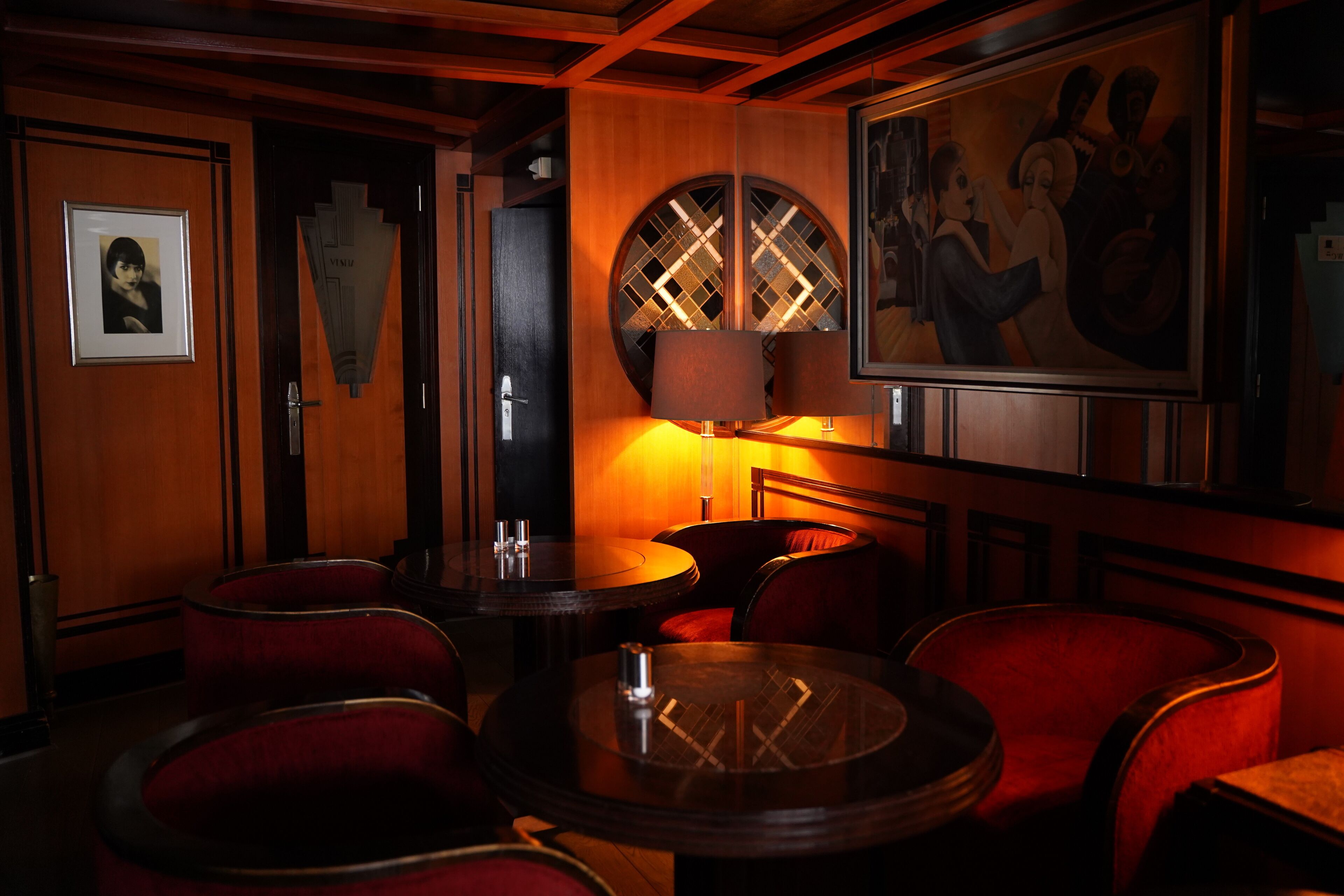 Un coin bar à la lumière tamisée, avec des boiseries, des chaises rembourrées de tissu rouge et une table ronde dont le plateau est en verre.
