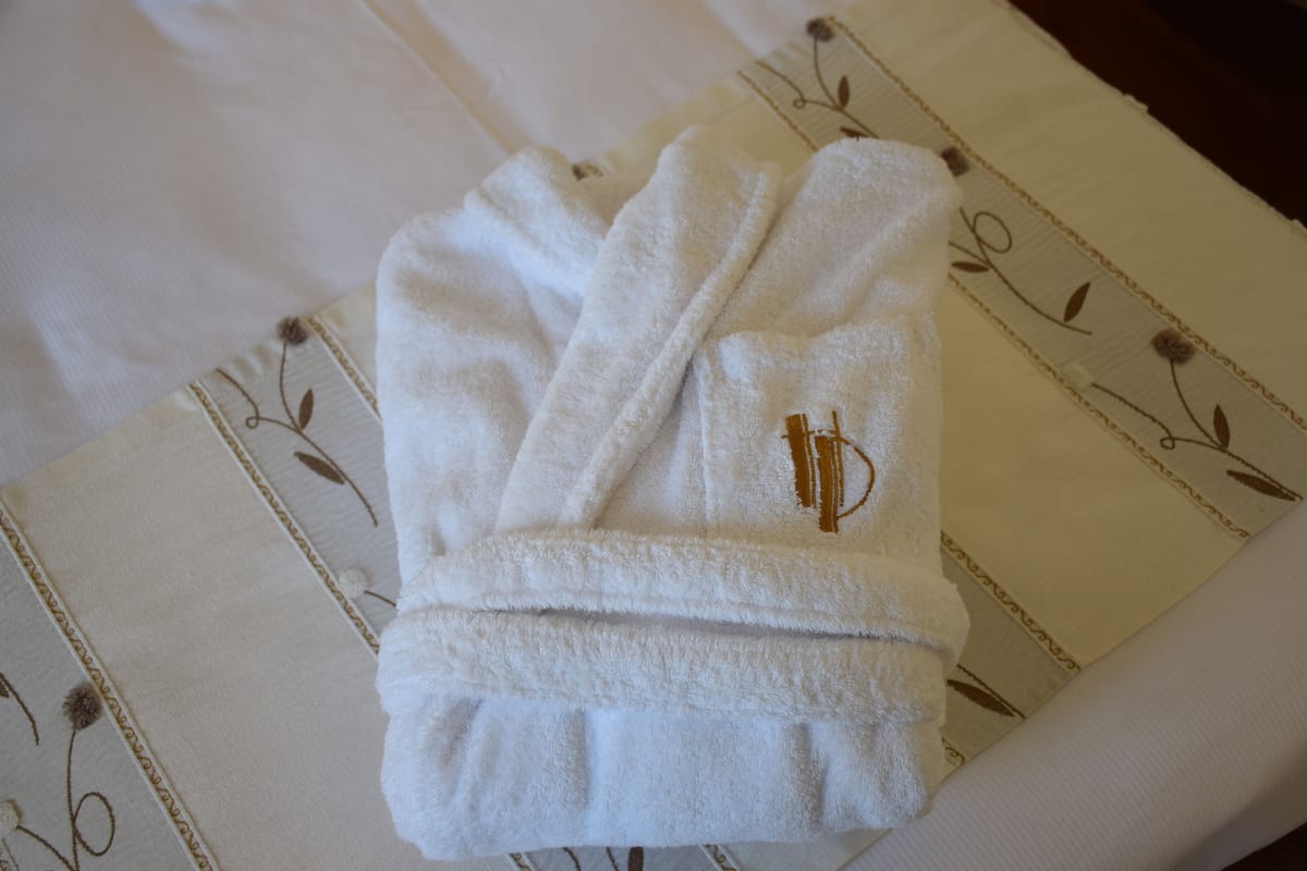 A neatly folded white bathrobe with a gold emblem on a bed with a floral-patterned pillow.