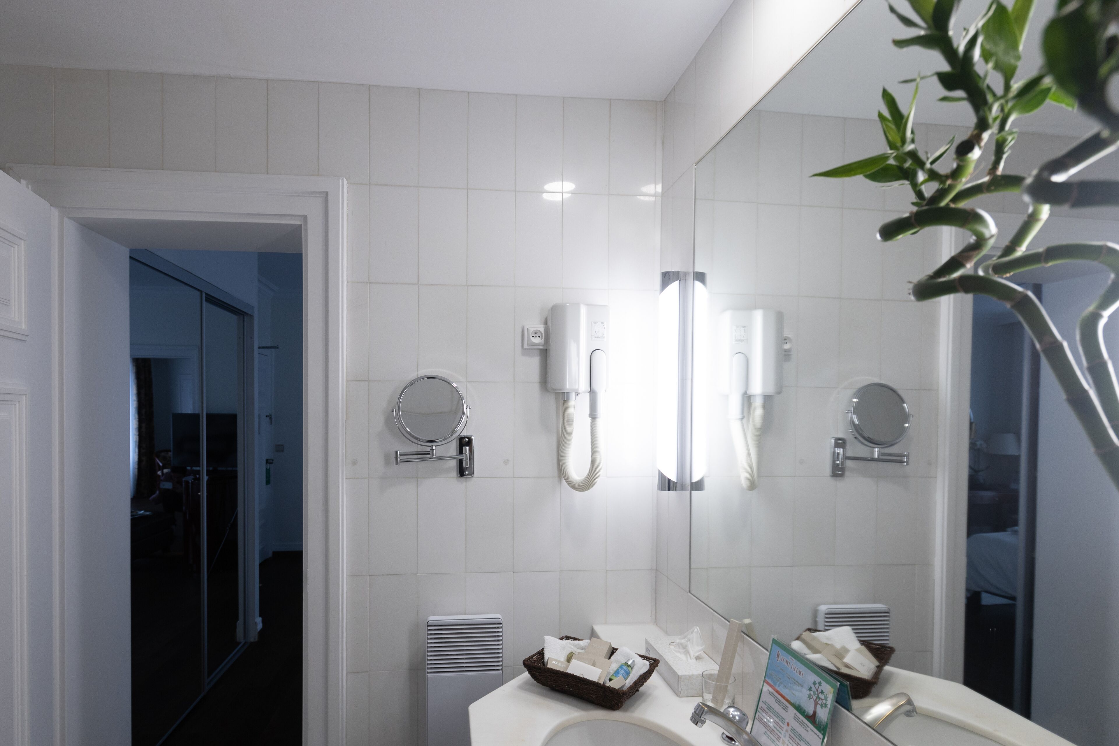 A modern bathroom with white tiles, a large mirror, two round mirrors, a sink with toiletries, and a heating vent.