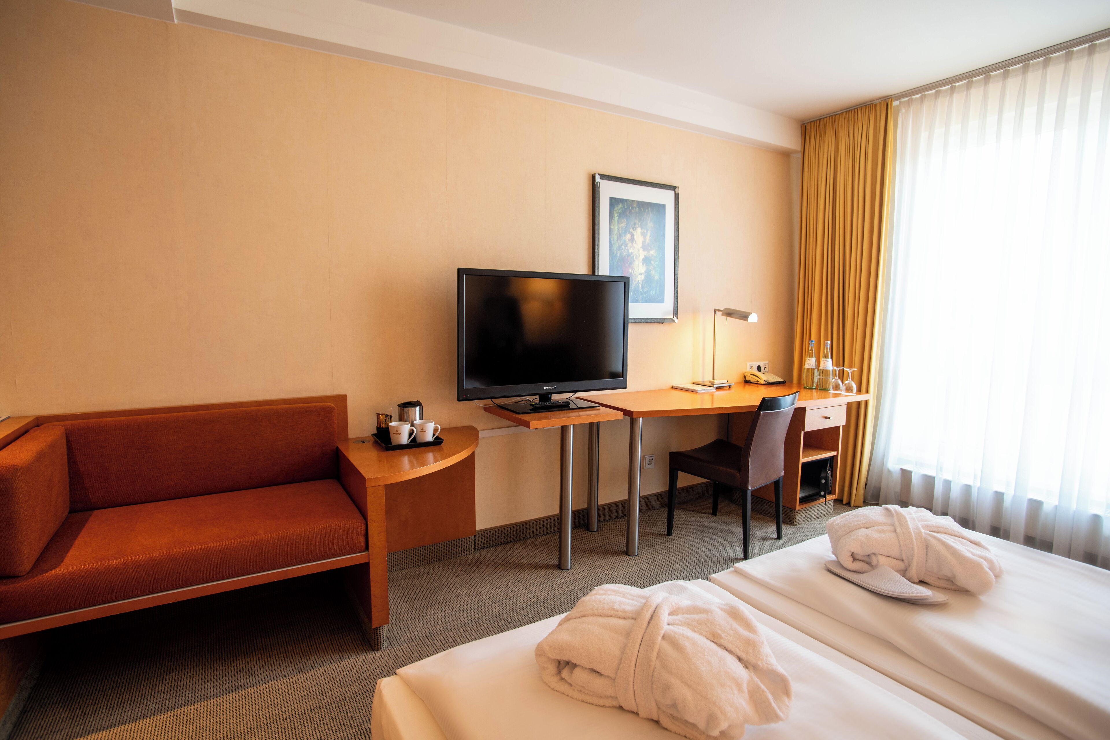 Ein Hotelzimmer mit Sofa, Fernseher, Schreibtisch mit Stuhl, Bett mit Handtüchern und Fenster mit Vorhängen.