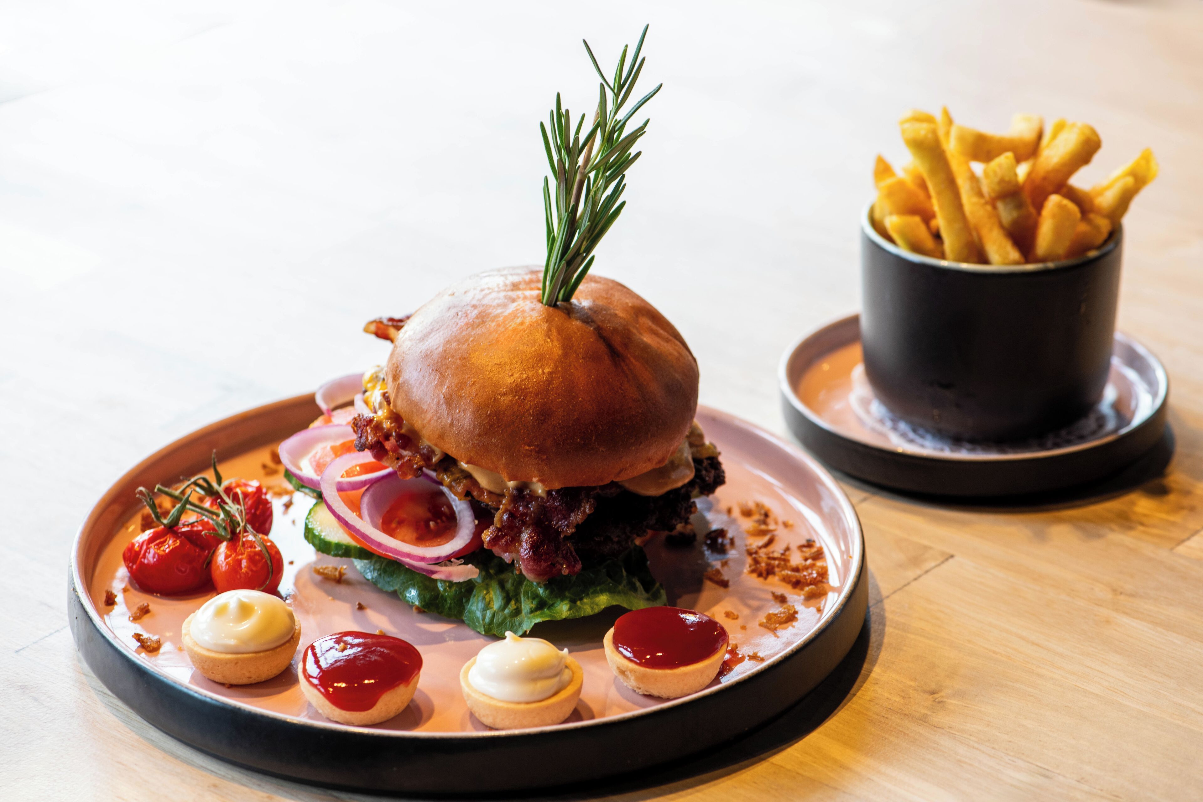 A burger with a sprig of rosemary on top, accompanied by cherry tomatoes, onions, and a small cup of fries.
