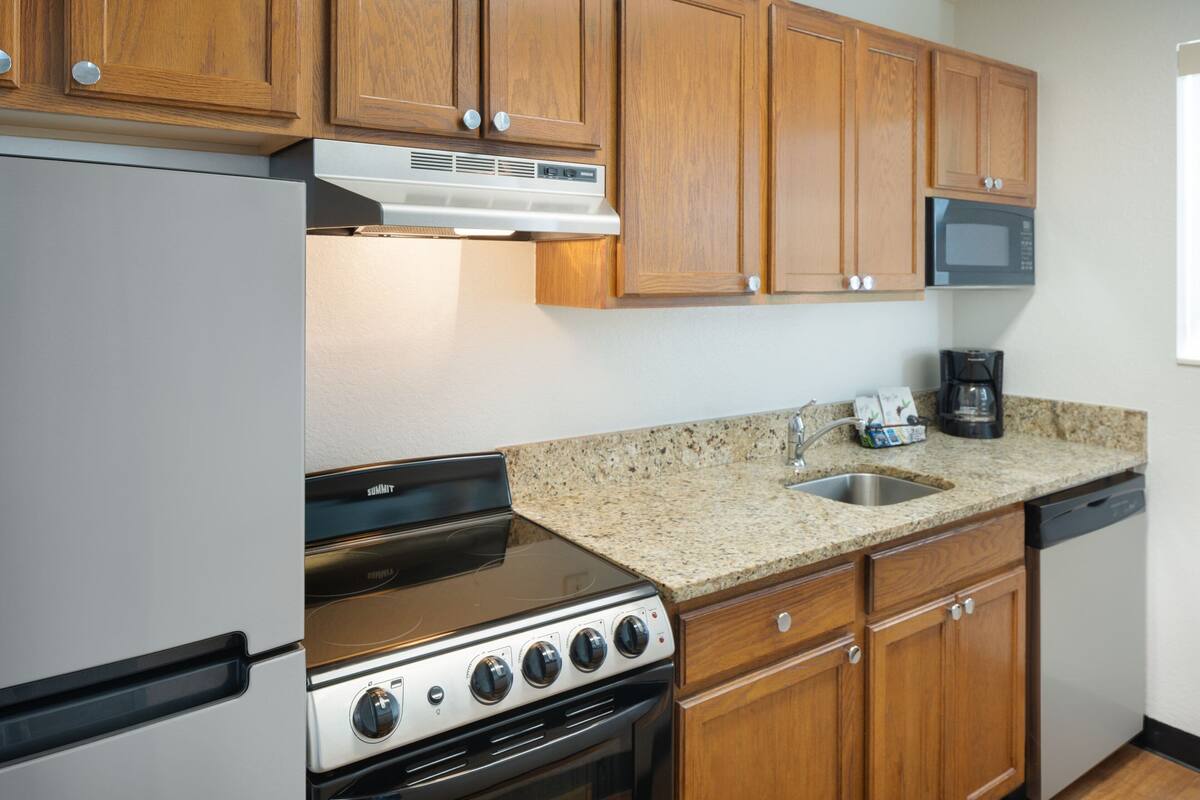 Una cocina con armarios de madera, un refrigerador de acero inoxidable, horno empotrado, microondas y encimera de granito.