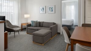 Una habitación de hotel moderna con un sofá gris, una mesita de madera, una lámpara pequeña y una cama con ropa blanca de cama.