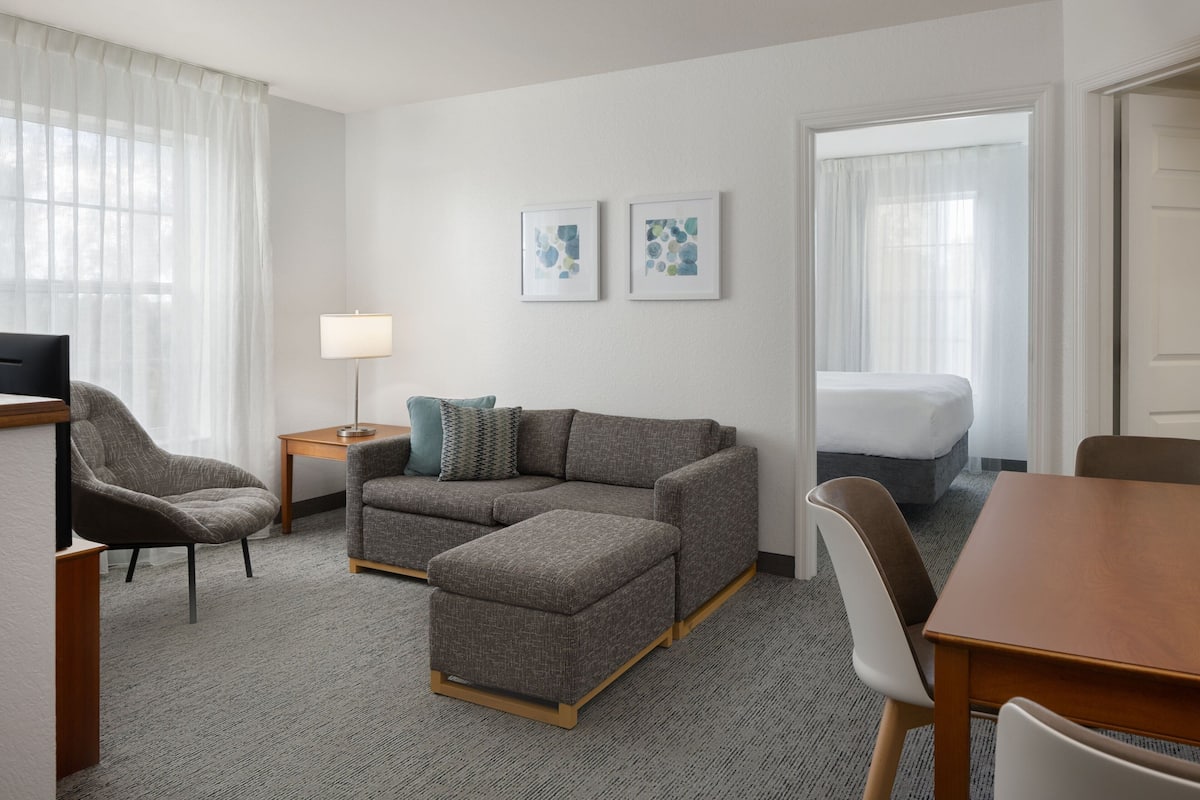 Una habitación de hotel moderna con un sofá gris, una mesita de madera, una lámpara pequeña y una cama con ropa blanca de cama.