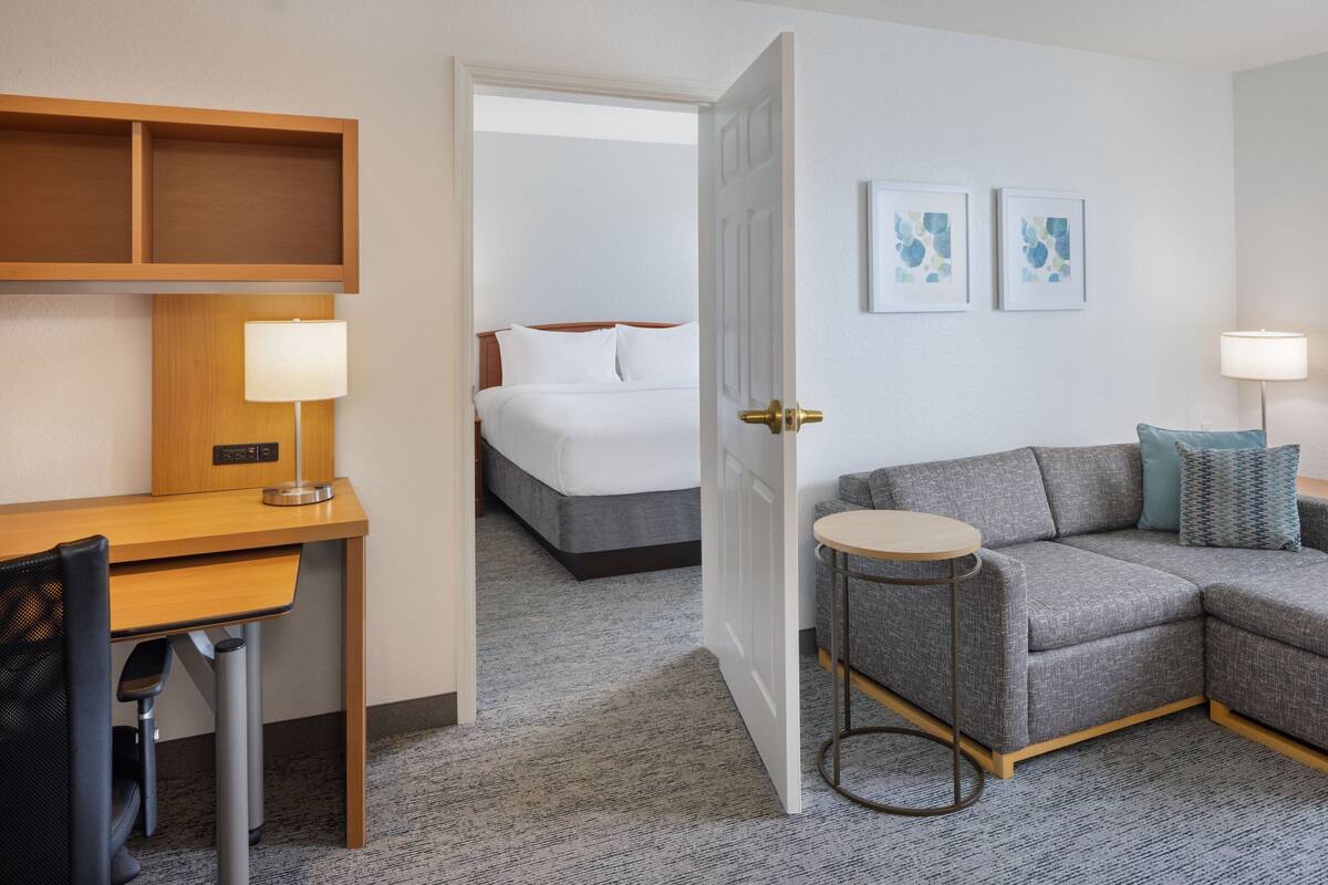 Una habitación de hotel con escritorio, sofá gris, cama y dos obras de arte colgadas en la pared.