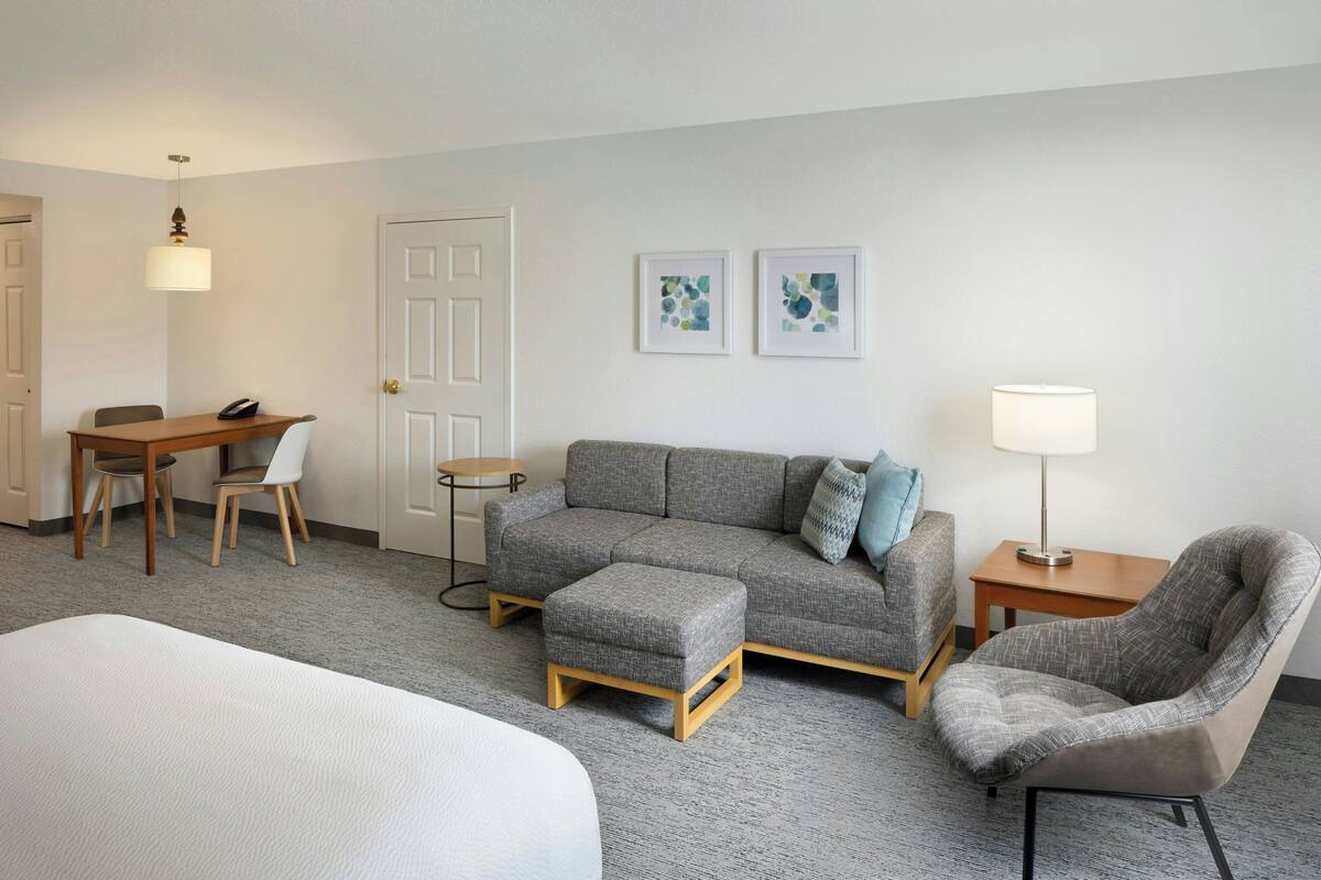 Habitación de hotel con sofá gris, sillón a juego, una mesita, un escritorio con silla y una cama.
