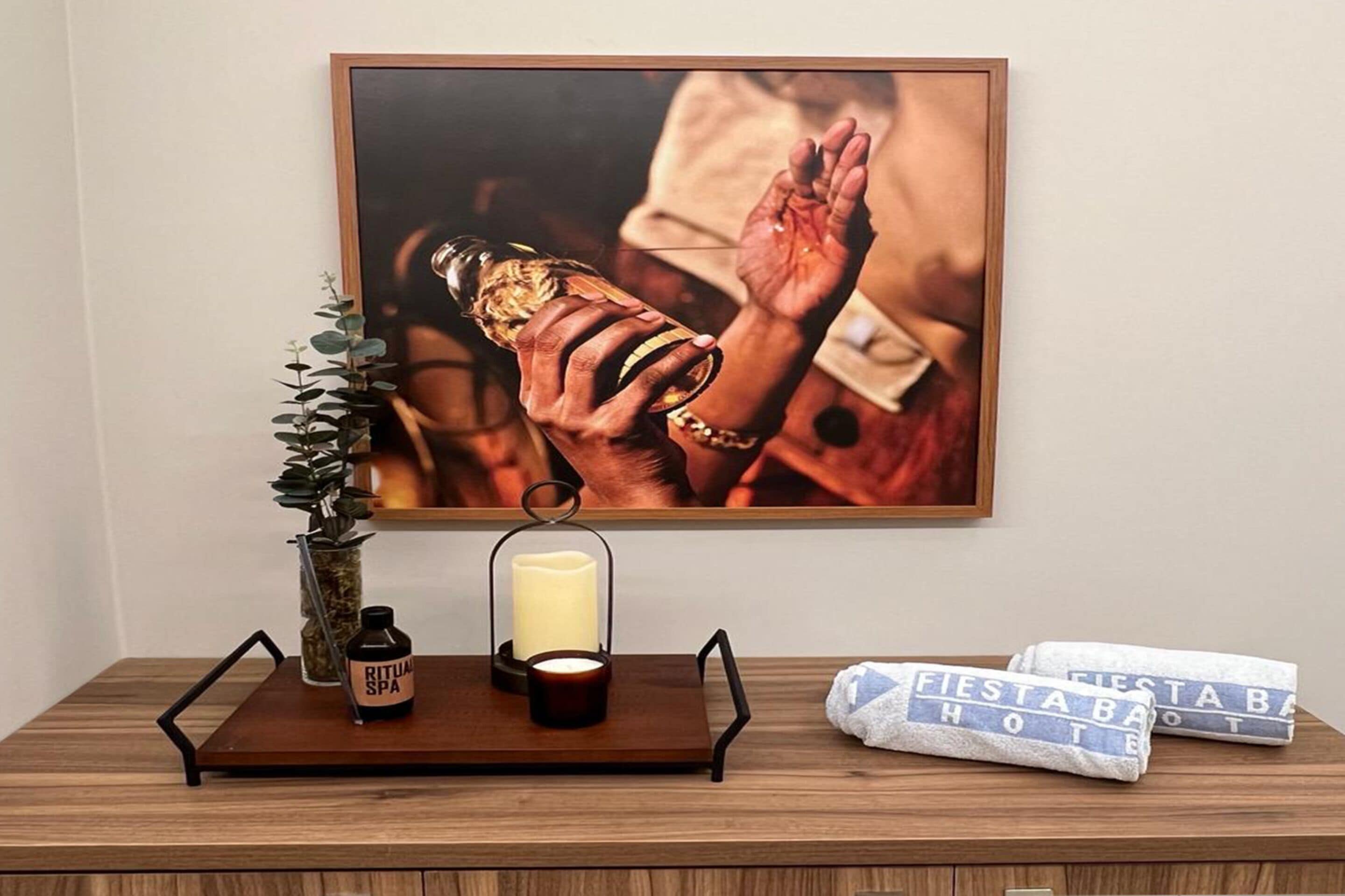 A framed artwork of hands holding a bottle, a candle, and a bottle labeled "RITUAL SPA" on a wooden table.