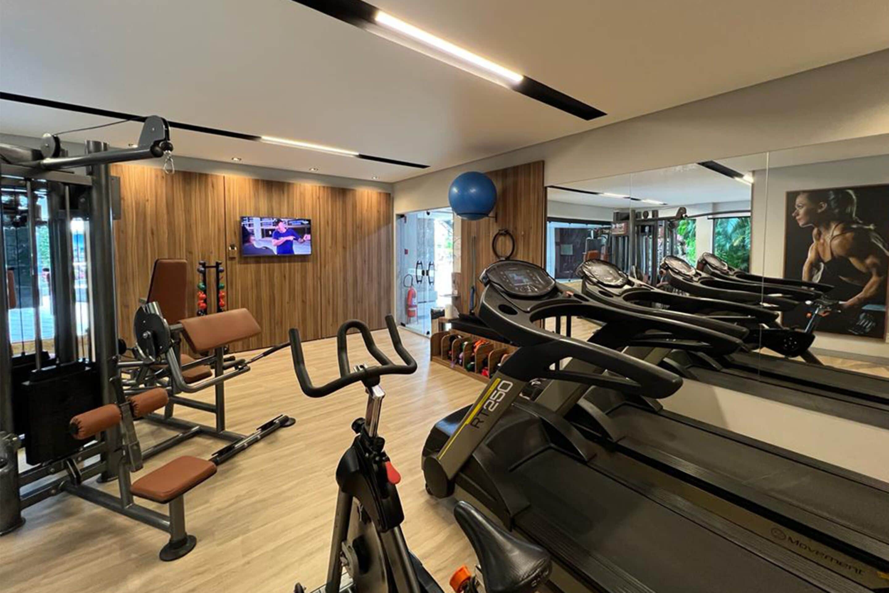 Uma academia com equipamentos de cardio, máquinas de peso e uma tela de televisão.