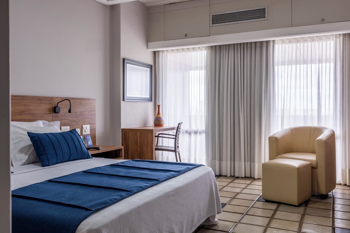 A hotel room with a bed, a desk, a chair, and a large window with curtains.