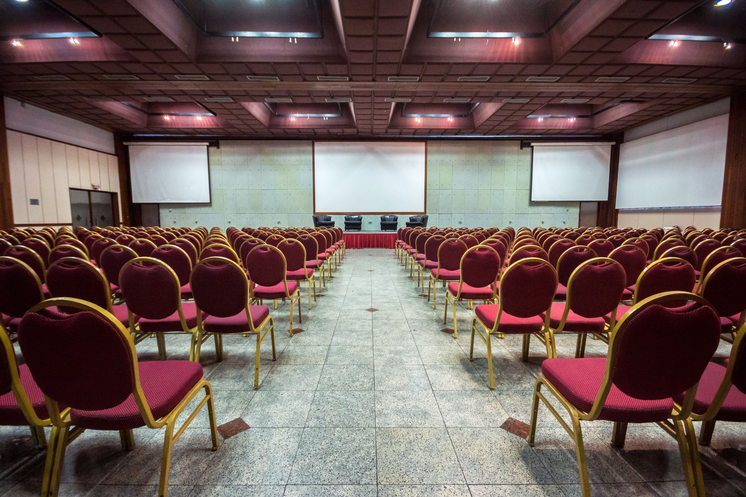 Uma sala de conferências com fileiras de cadeiras vermelhas e uma tela grande na frente.