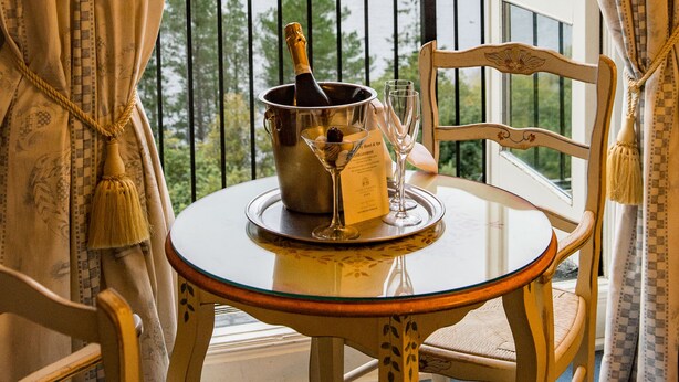 Una mesa con una botella de champán, una copa y un menú en un balcón con vista a un lago y montañas al fondo.
