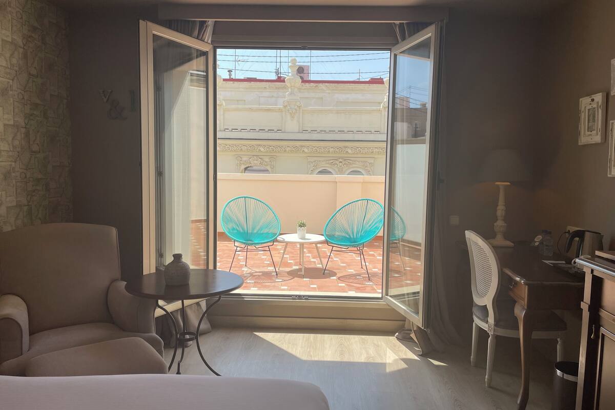 A hotel room with a bed, a desk, and a balcony with two chairs.