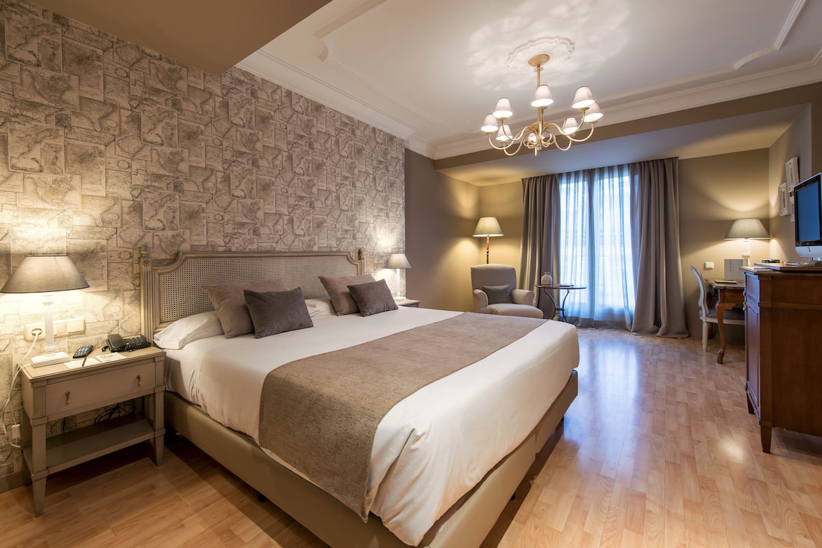 A hotel room with a large bed, a desk, a chair, a television, and a chandelier.