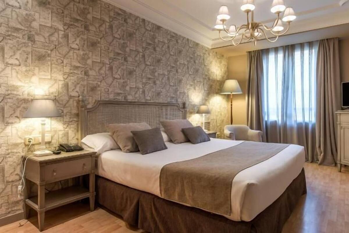 A hotel room with a stone-patterned wall, a large bed with multiple pillows, bedside lamps, a TV on a cabinet, and a chandelier.