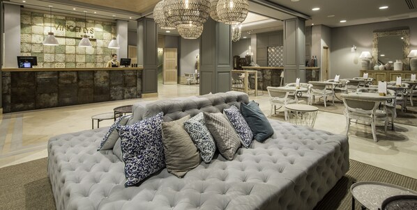 Een hotel lobby met een receptiebalie, een gestoffeerde bank met gekleurde kussens, een kroonluchter en een eetgedeelte met witte stoelen en tafels.