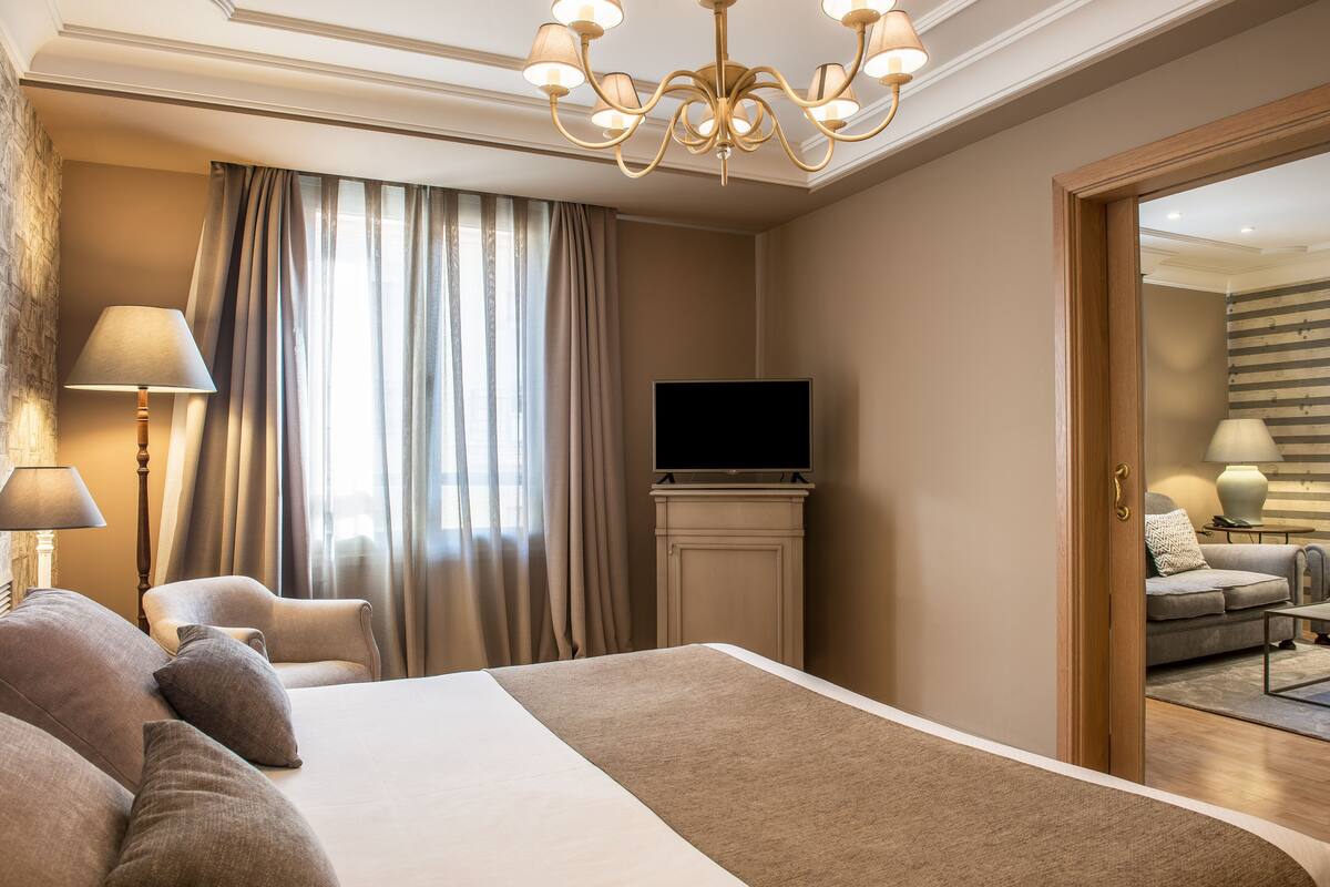 A hotel room with a large bed, a flat-screen TV, a chandelier, and a view of the adjacent living area.