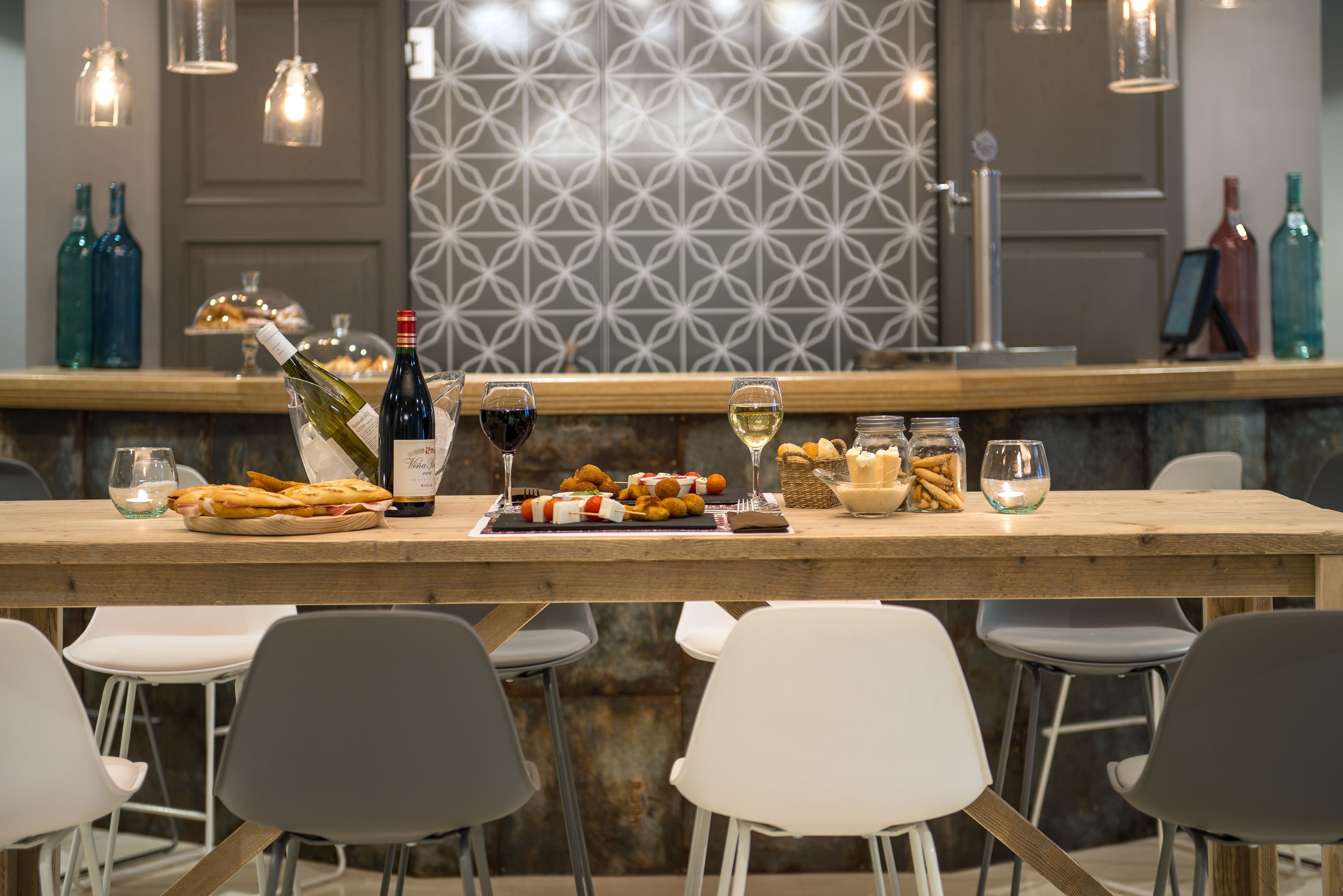 Una zona de bar con mostrador de madera, sillas blancas y grises, y botellas de vino.