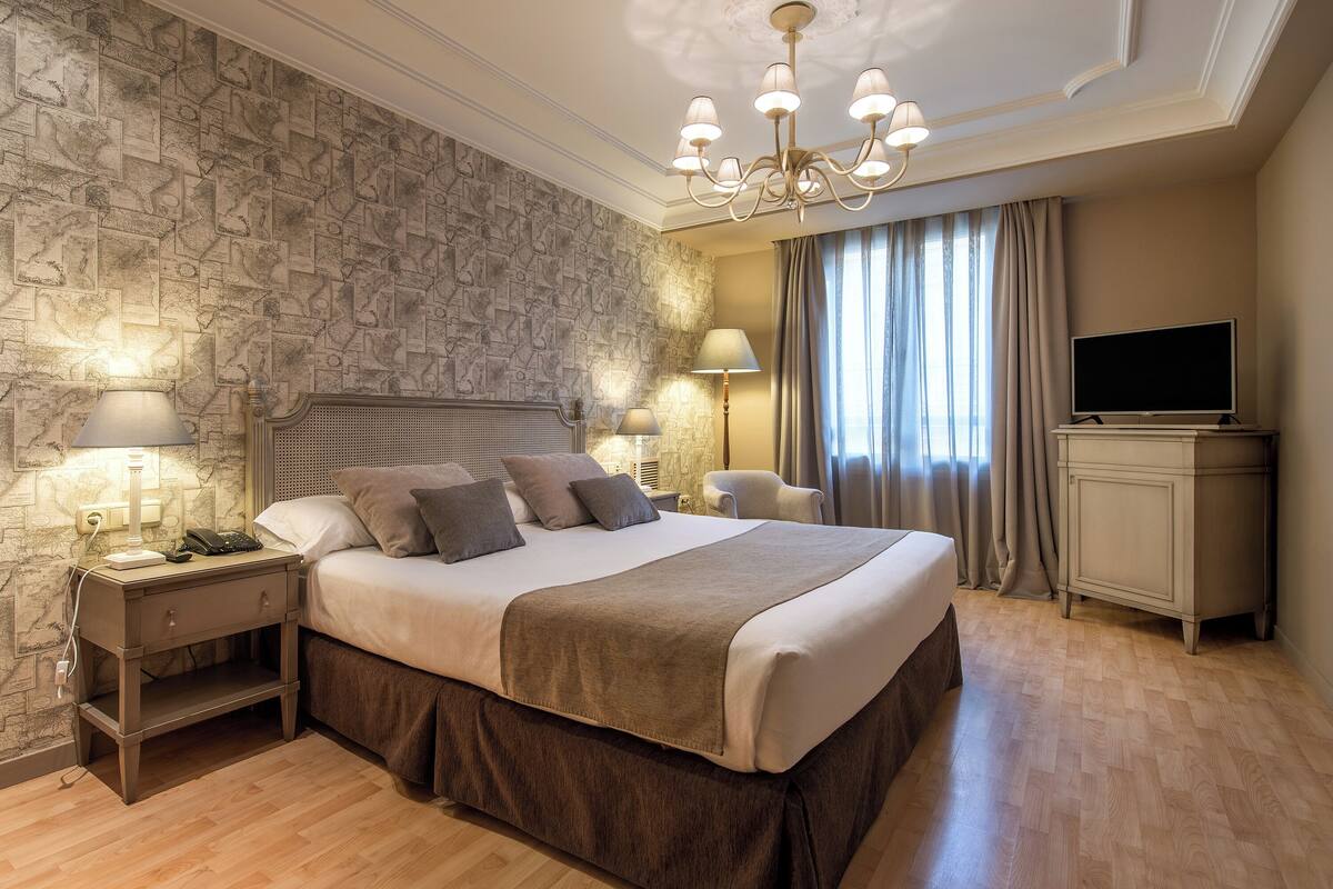 A bedroom with a large bed, a nightstand with a lamp, a television, and a stone-patterned wall.