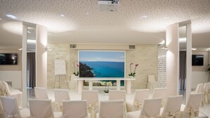 A conference room with a large screen displaying a scenic view, white chairs with bows, and a whiteboard on the wall. - Scoglio della Galea Village & Spa (Ricadi)