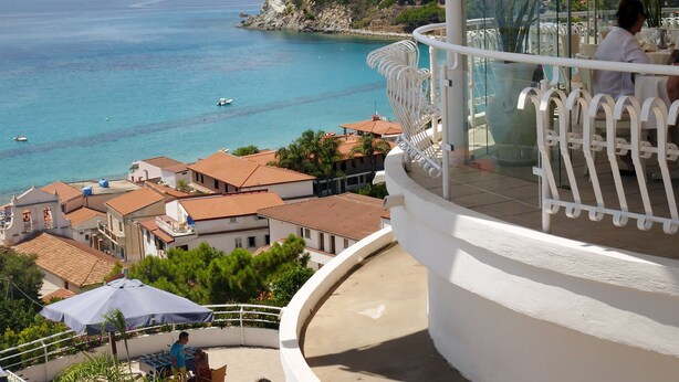 A balcony with a view of the sea, a dining area with tables and chairs, and a building with a tiled roof. - Scoglio della Galea Village & Spa (Ricadi)