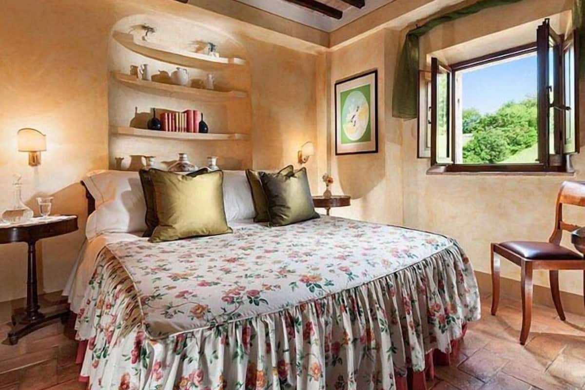 A bedroom with a bed, a nightstand, a chair, a window, and a shelf with books and decorative items.