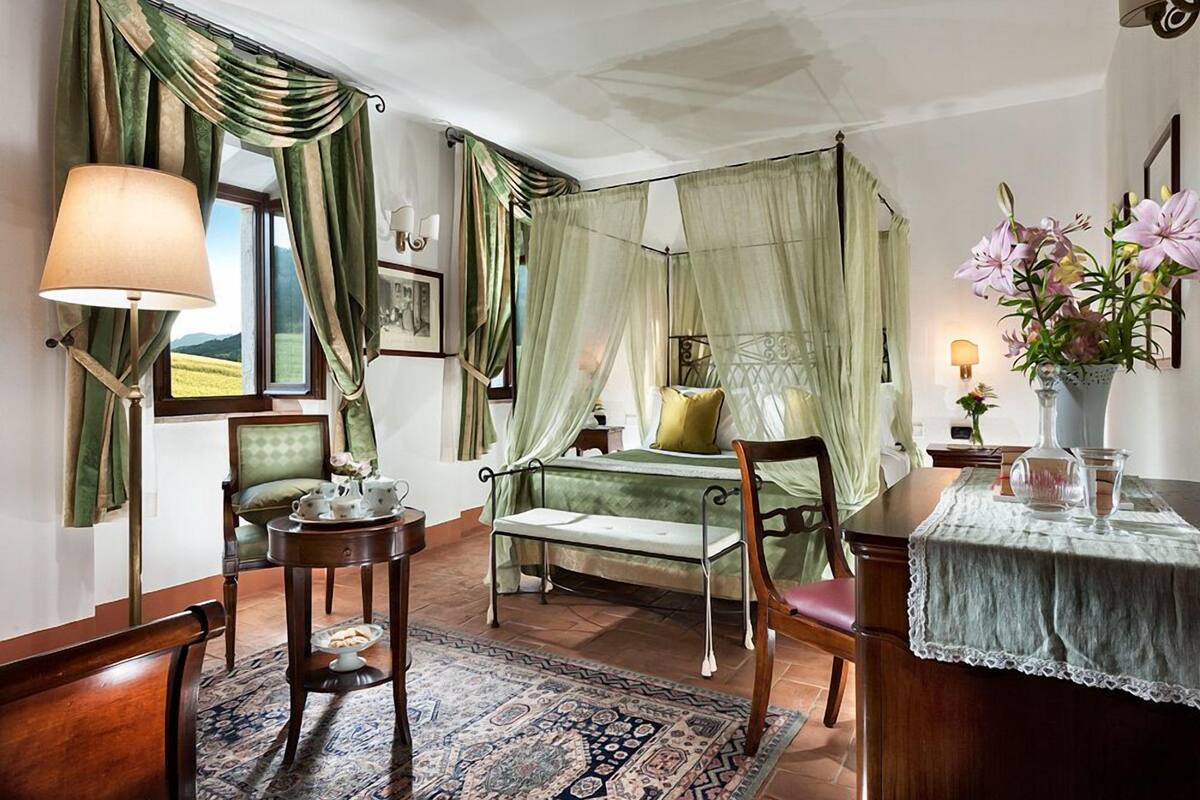 A bedroom with a canopy bed, a bedside table with a lamp, a chair, a small table with a vase of flowers, and a view of the outdoors.