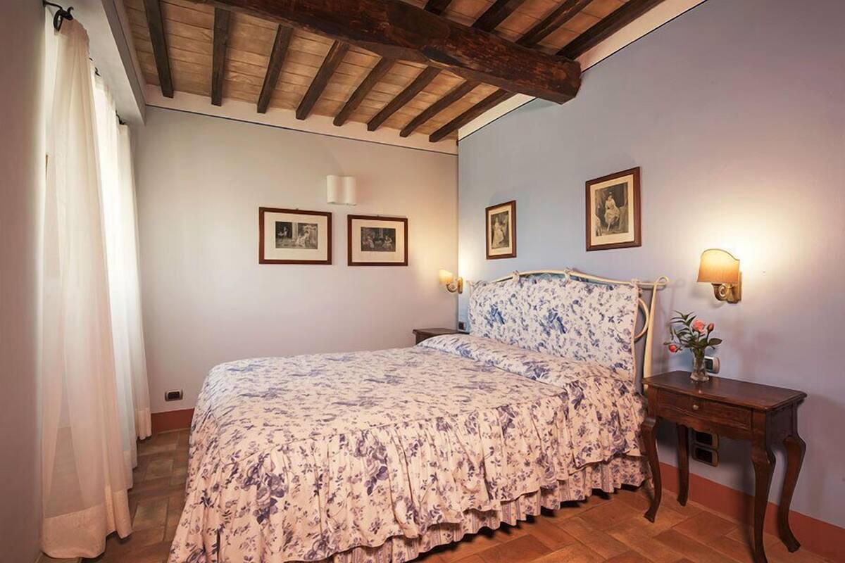 A bedroom with a bed, wooden ceiling beams, a nightstand with a lamp, and framed pictures on the wall.