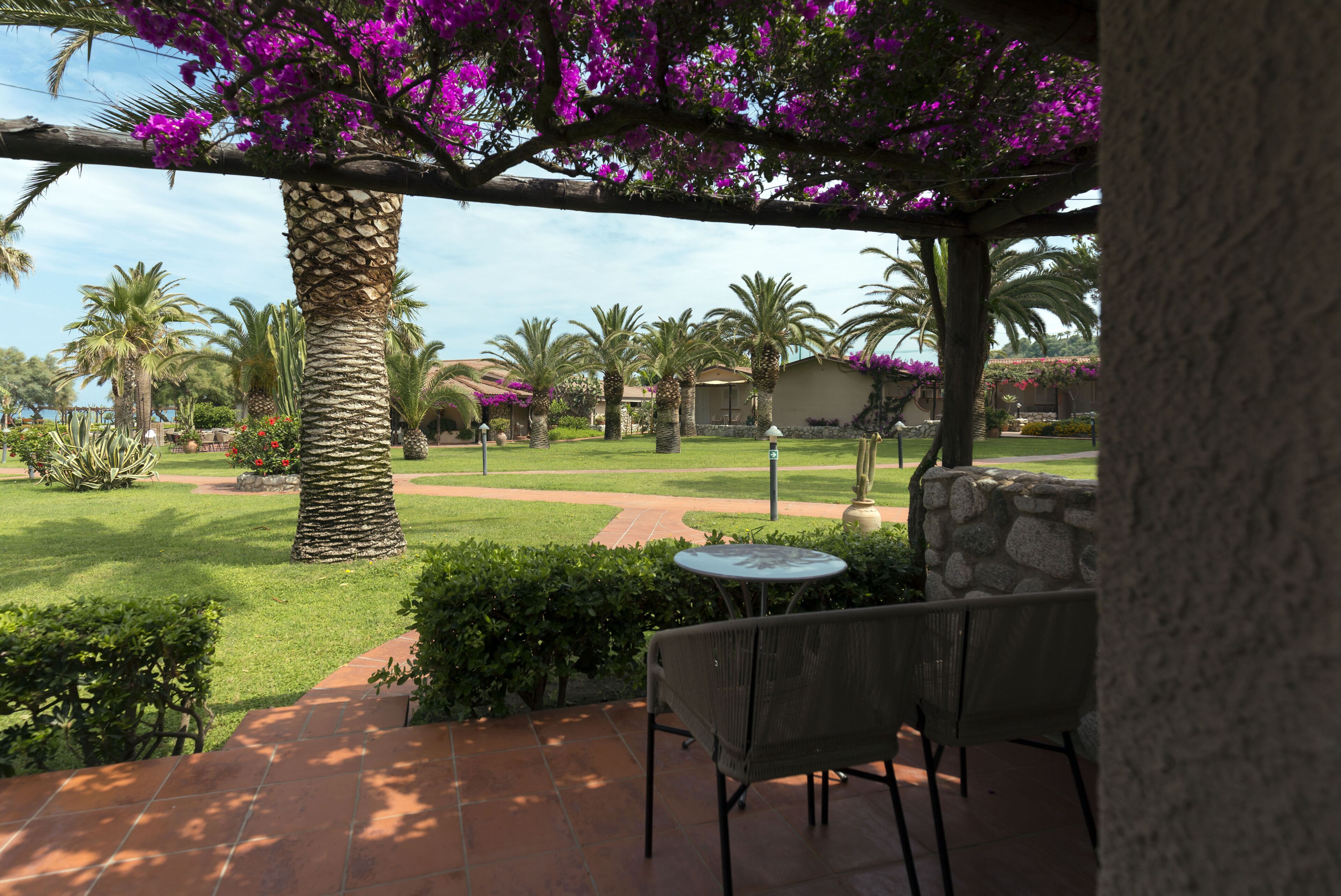 Un espace patio agrémenté d’un mur en pierre, d’une table et de chaises, donnant sur un jardin bien entretenu avec des palmiers et des fleurs violettes.