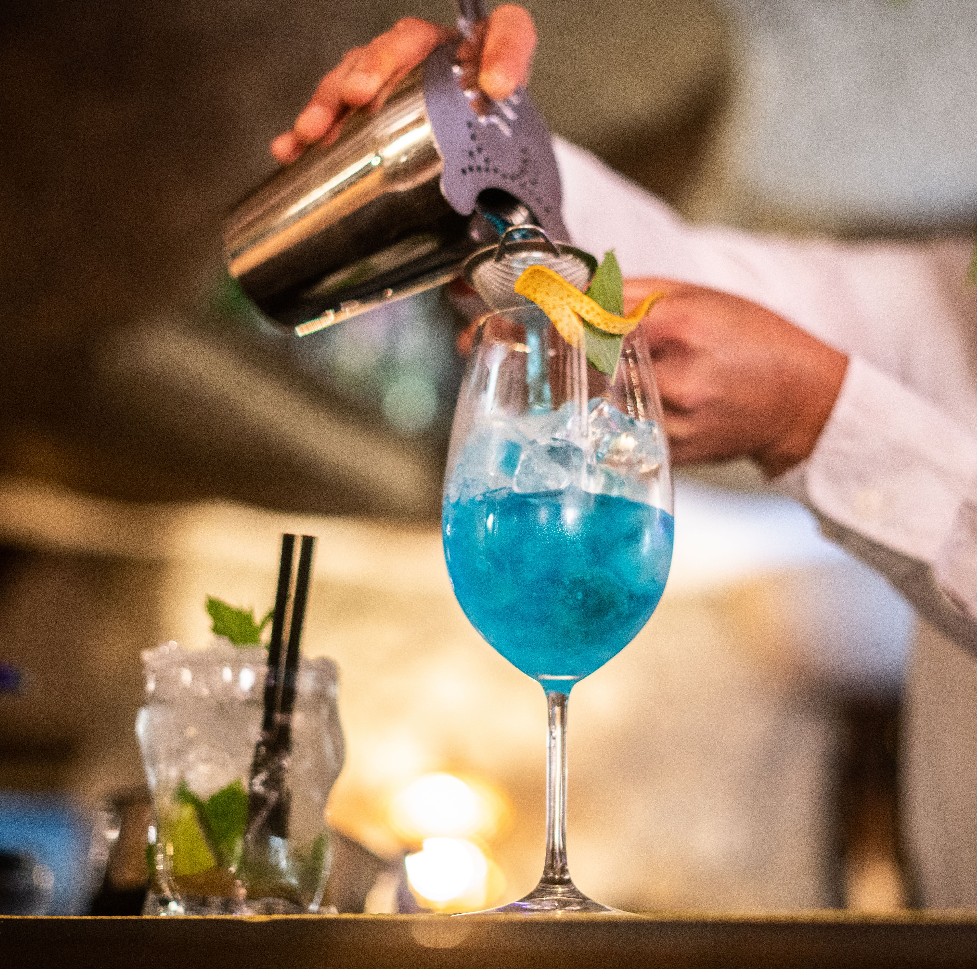 A bartender pouring a blue cocktail into a glass with ice and garnished with a lemon twist.