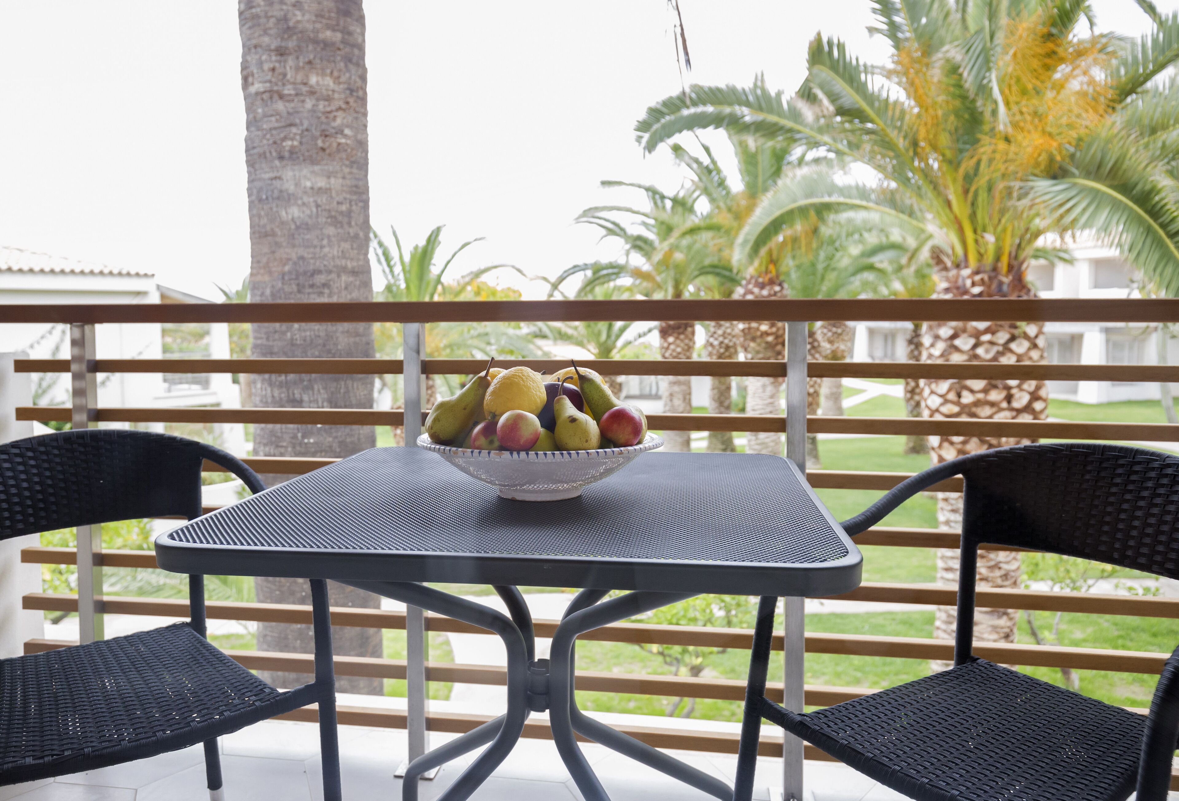 A table with a bowl of fruit on a balcony with wicker chairs and palm trees in the background.