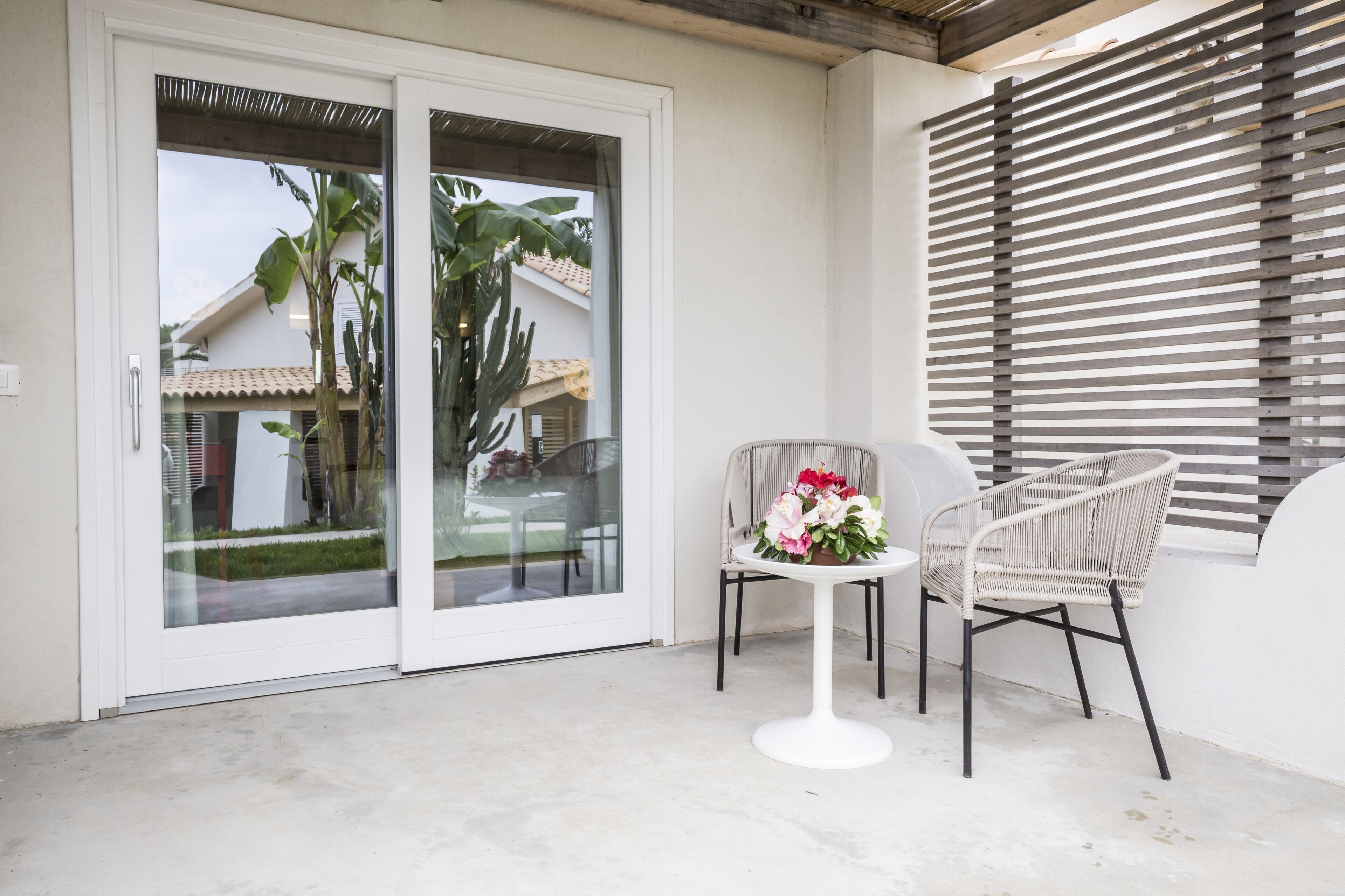Un balcón con una mesa blanca y dos sillas de mimbre, un ventanal de vidrio y una ventana con persianas horizontales.