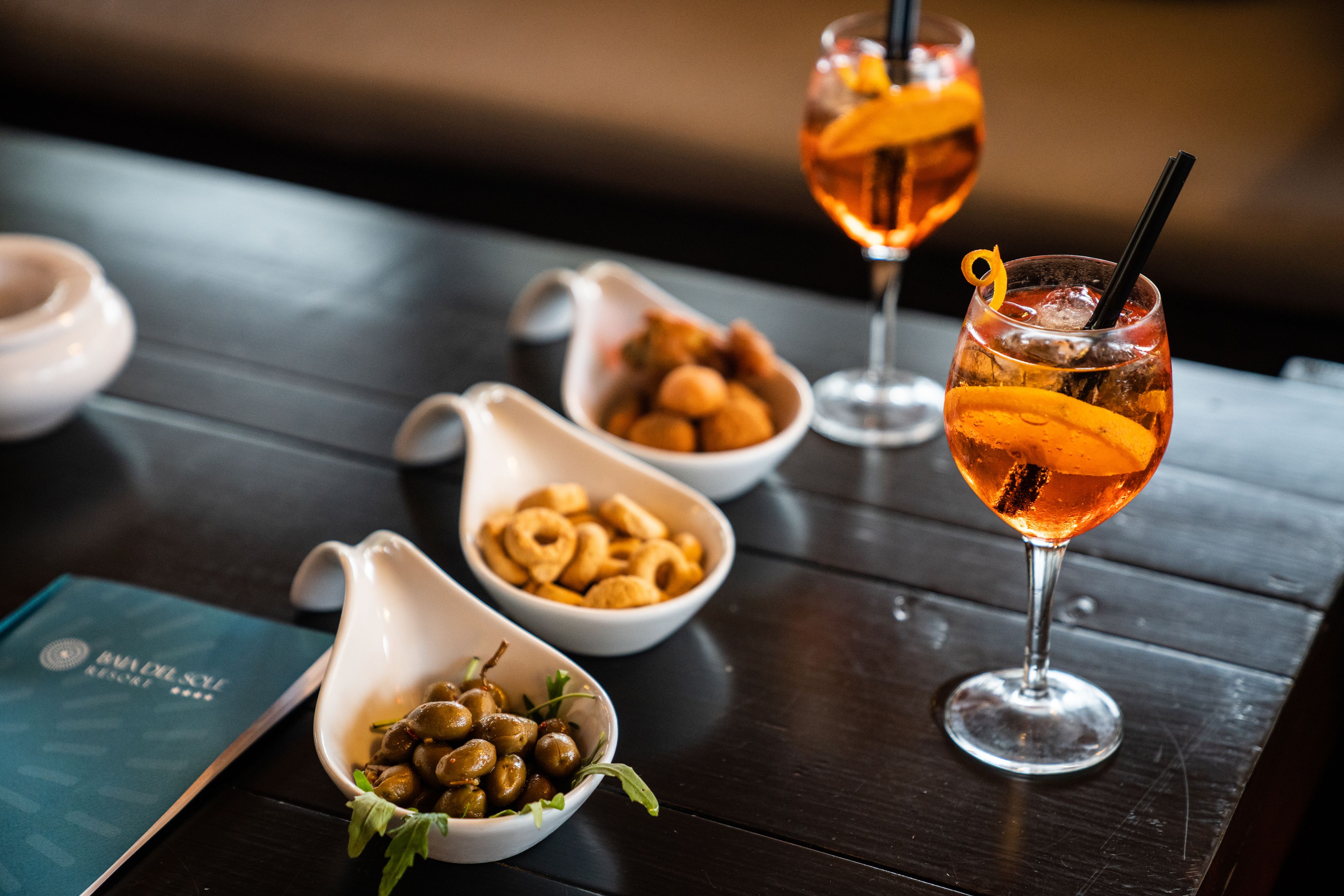A table with two glasses of amber-colored cocktails, a bowl of olives, a bowl of ring-shaped snacks, and a menu.