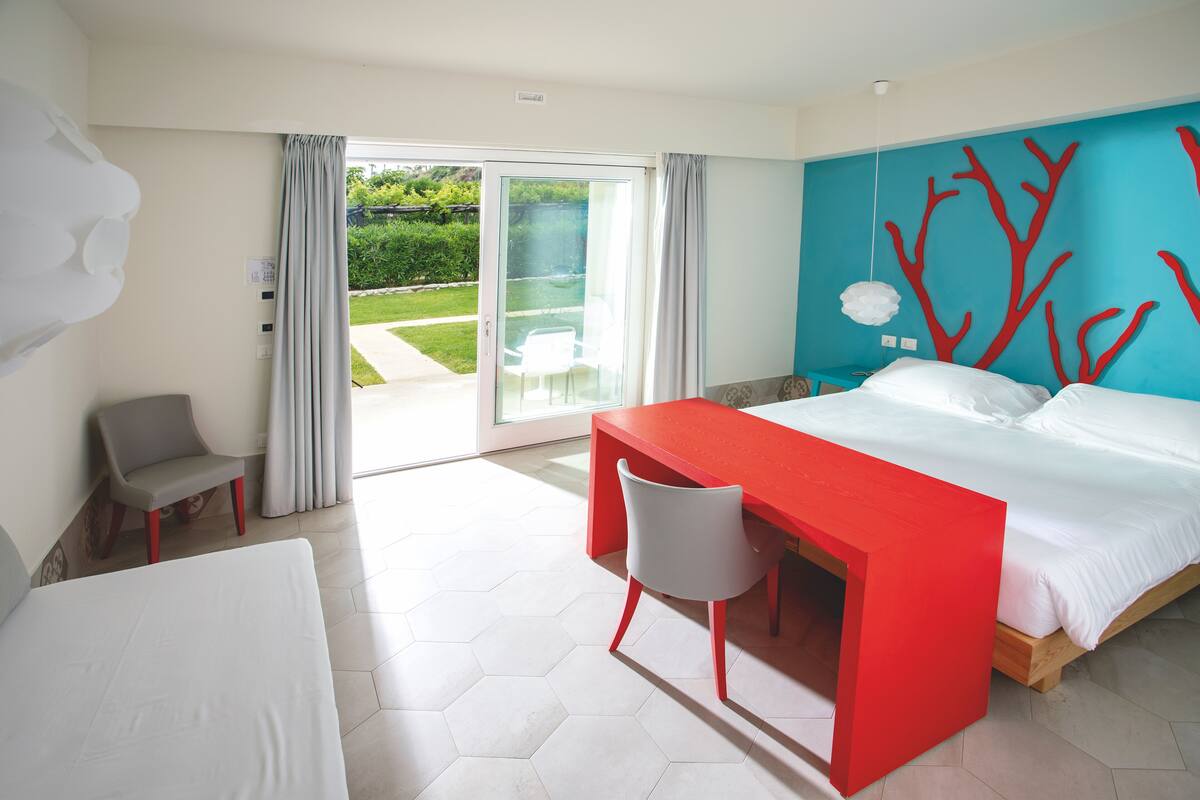 A modern hotel room with a large bed, a desk, and a chair. There is a sliding glass door leading to a balcony with a view of greenery.