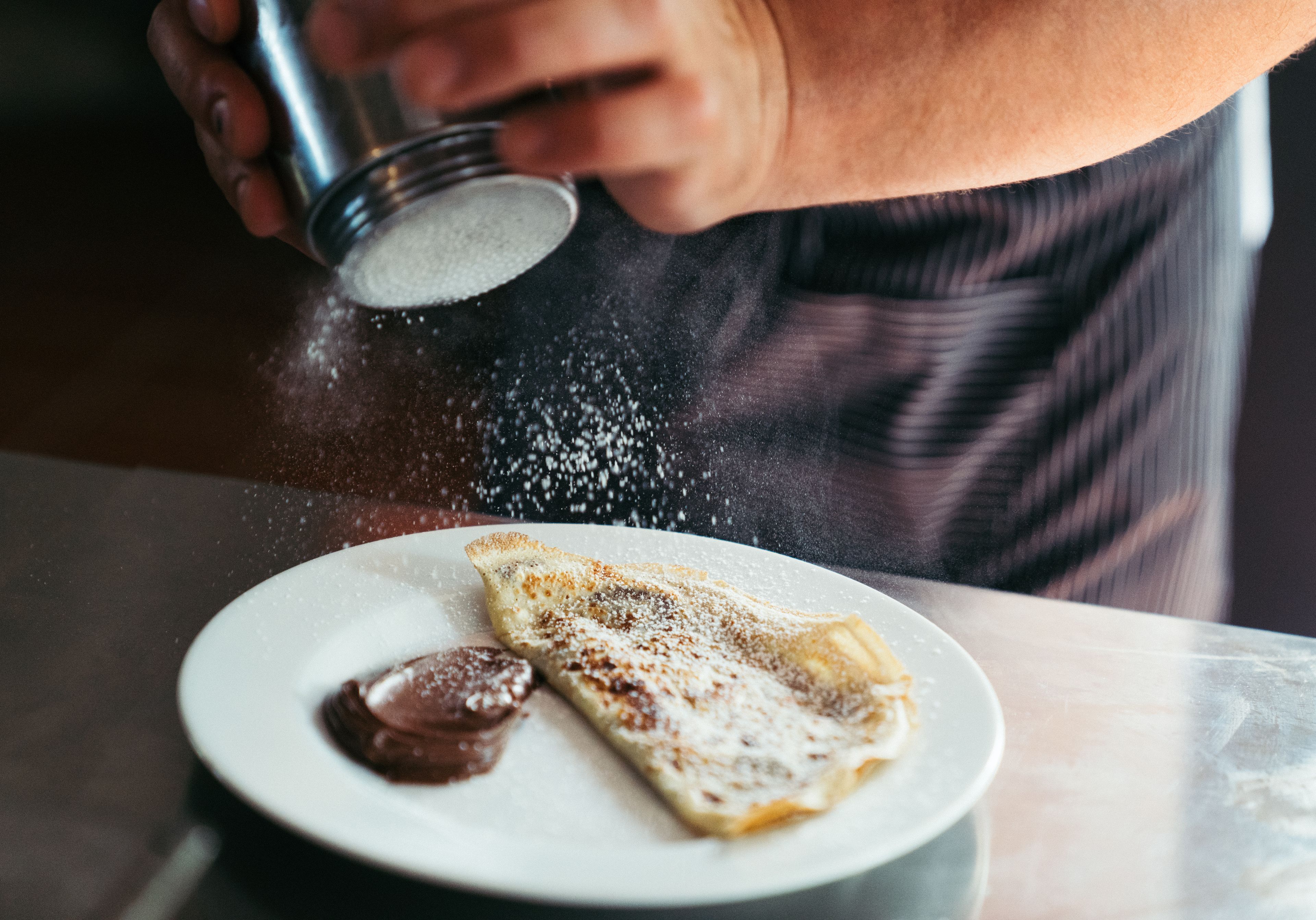 A person sprinkling powdered sugar on a crepe with chocolate sauce on a plate.
