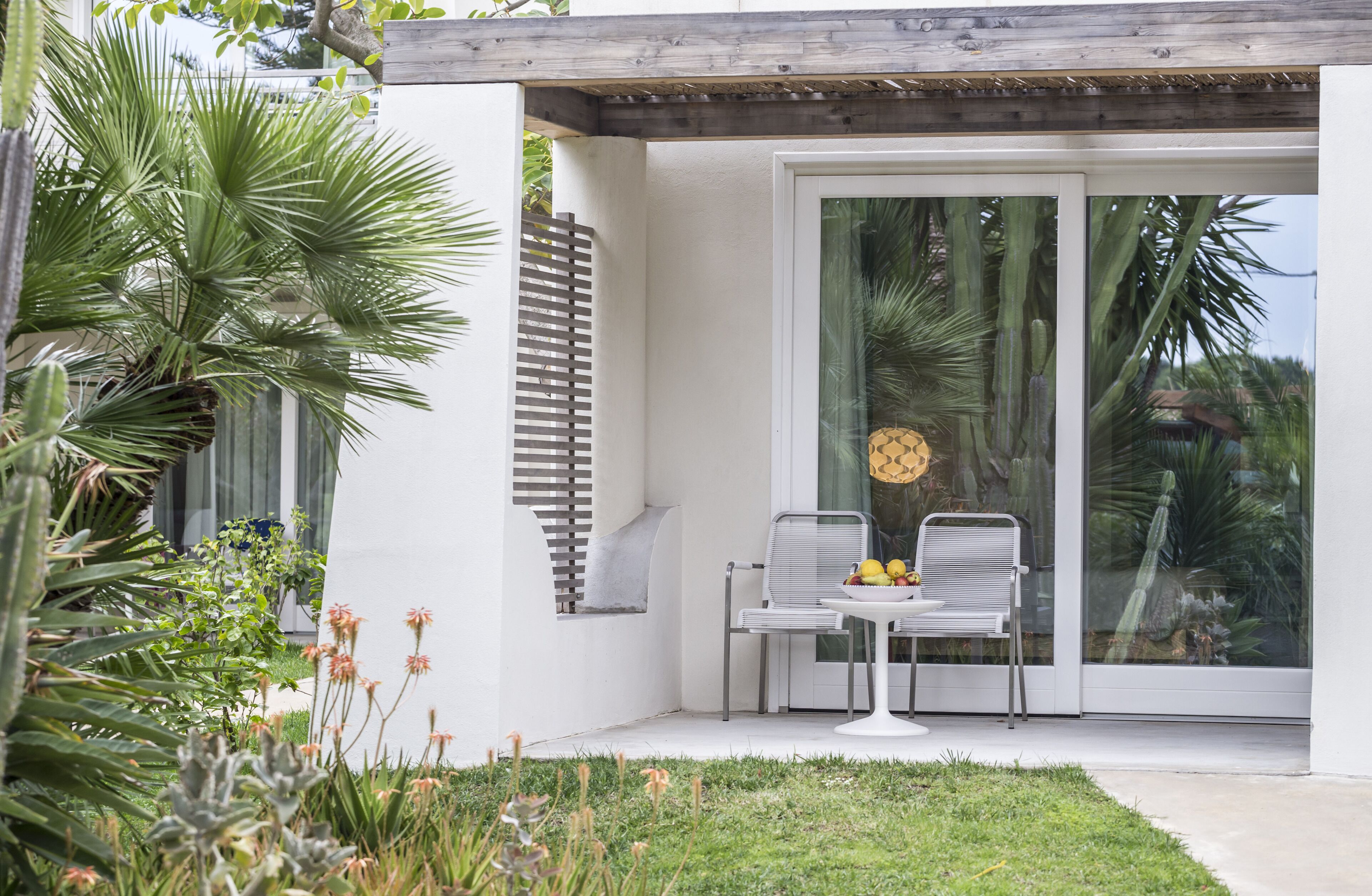 Une terrasse moderne avec une table blanche et deux chaises, entourée d’une végétation luxuriante et dotée d’une grande porte vitrée donnant sur un espace extérieur privé.