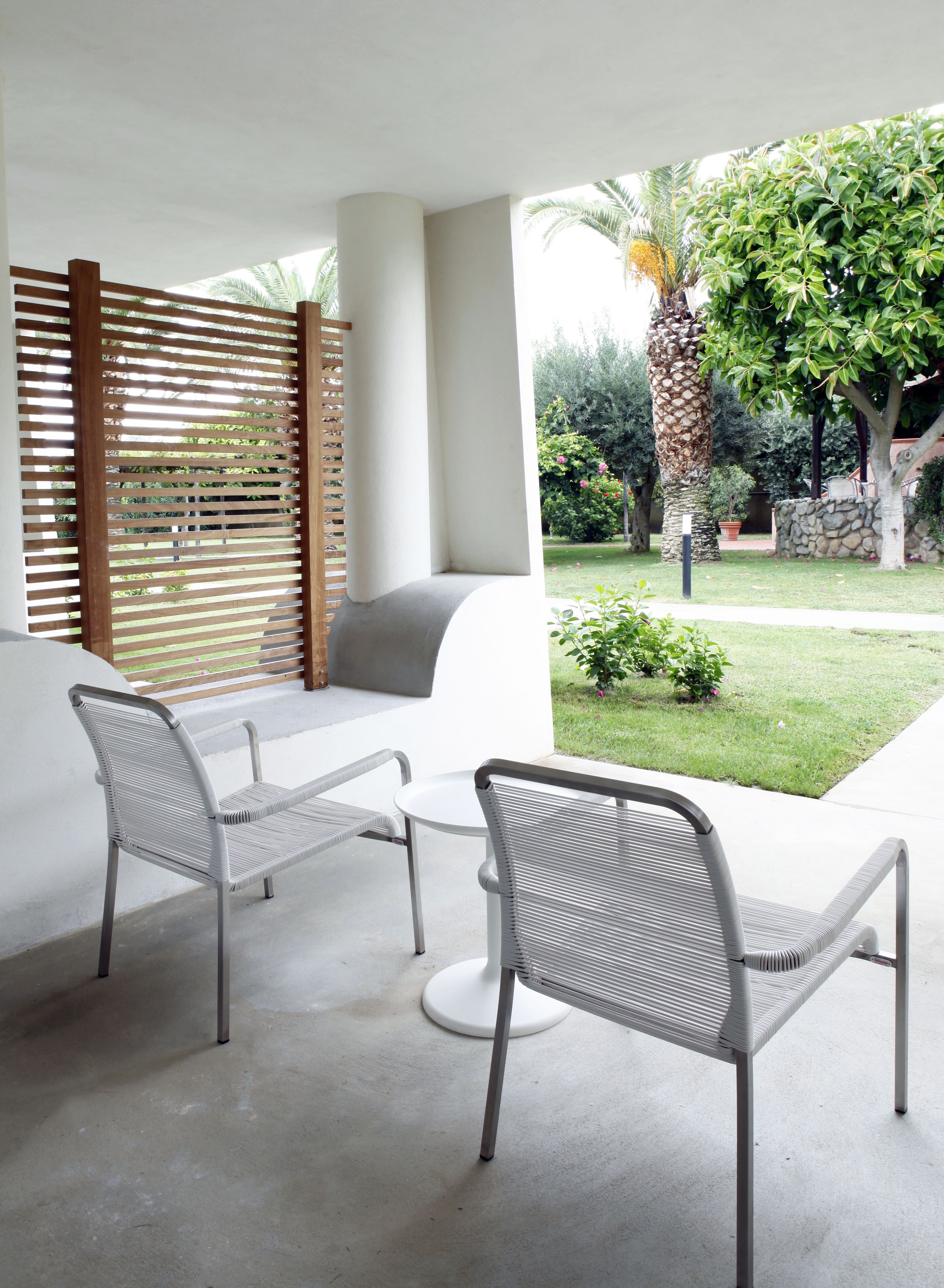 Un'area salotto esterna moderna con due sedie e un tavolo, un pannello divisorio in legno a listelli e vista su un giardino curato con diverse piante e una palma.
