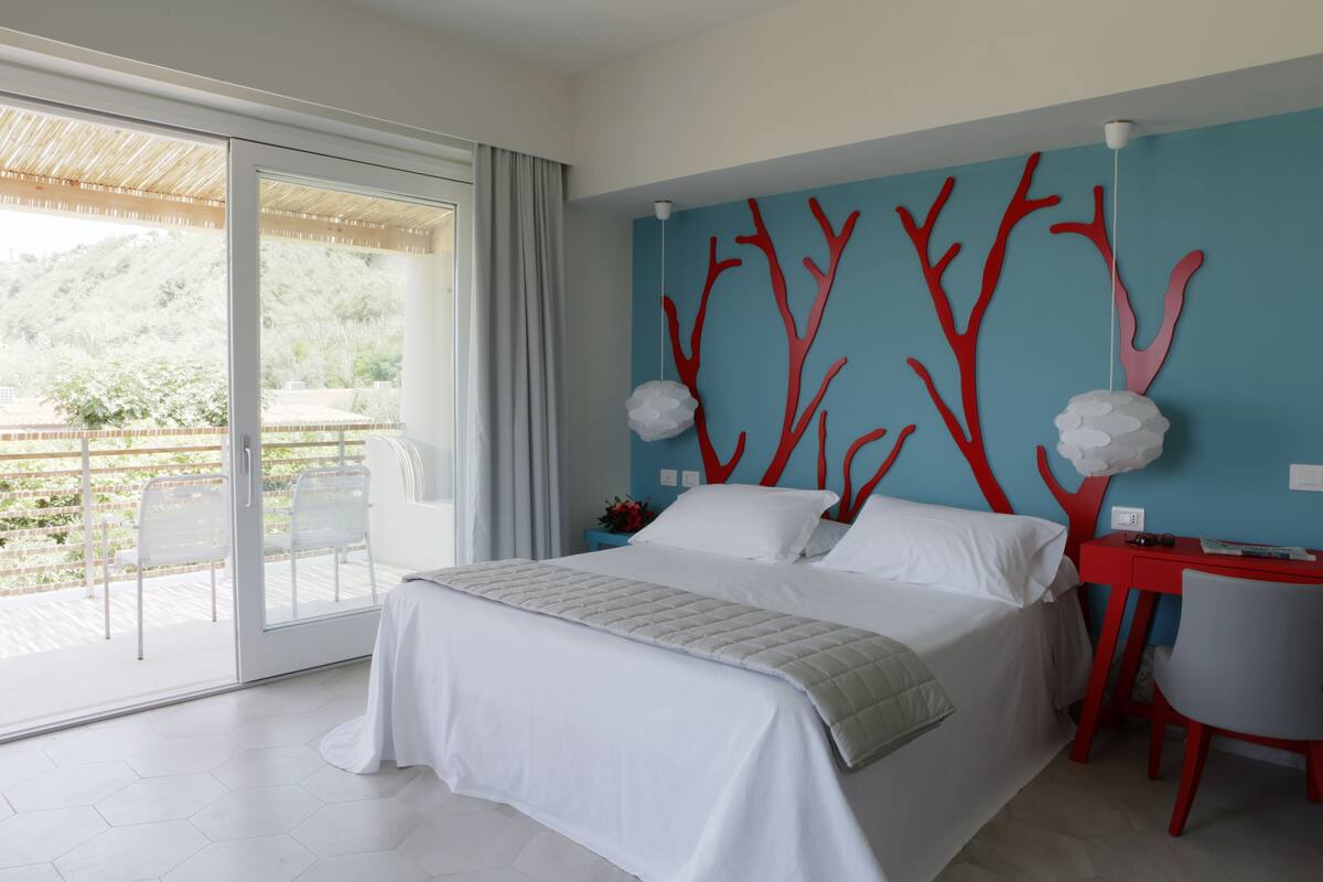 A modern bedroom with a bed, a desk, and a chair. There is a balcony with a view of greenery.