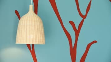 En hängande lampa med en vävd struktur, placerad framför en blå vägg med röda, abstrakta grenar.