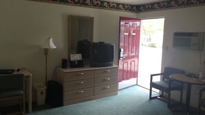 A hotel room with a television on a dresser, a desk with a lamp, a chair, a small trash bin, and a red door. - Prestige Inn West Lafayette (West Lafayette)