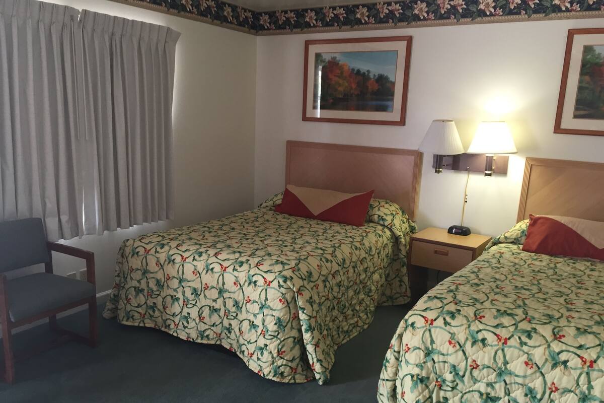 Ett hotellrum med två sängar, en stol, ett nattduksbord med en lampa och ett fönster med gardiner.