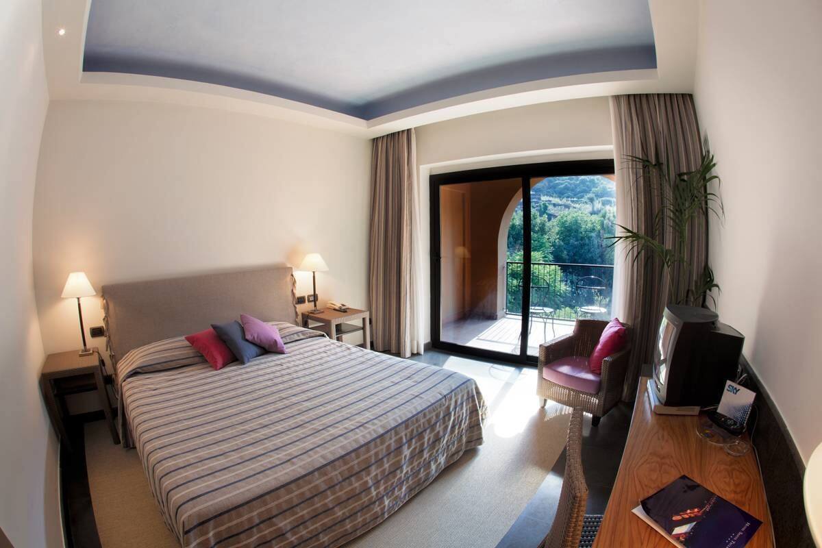 Ein Hotelzimmer mit einem großen Bett, zwei Nachttischen mit Lampen, einem Fernseher, einem Schreibtisch mit Stuhl und einem Balkon mit Blick ins Grüne.