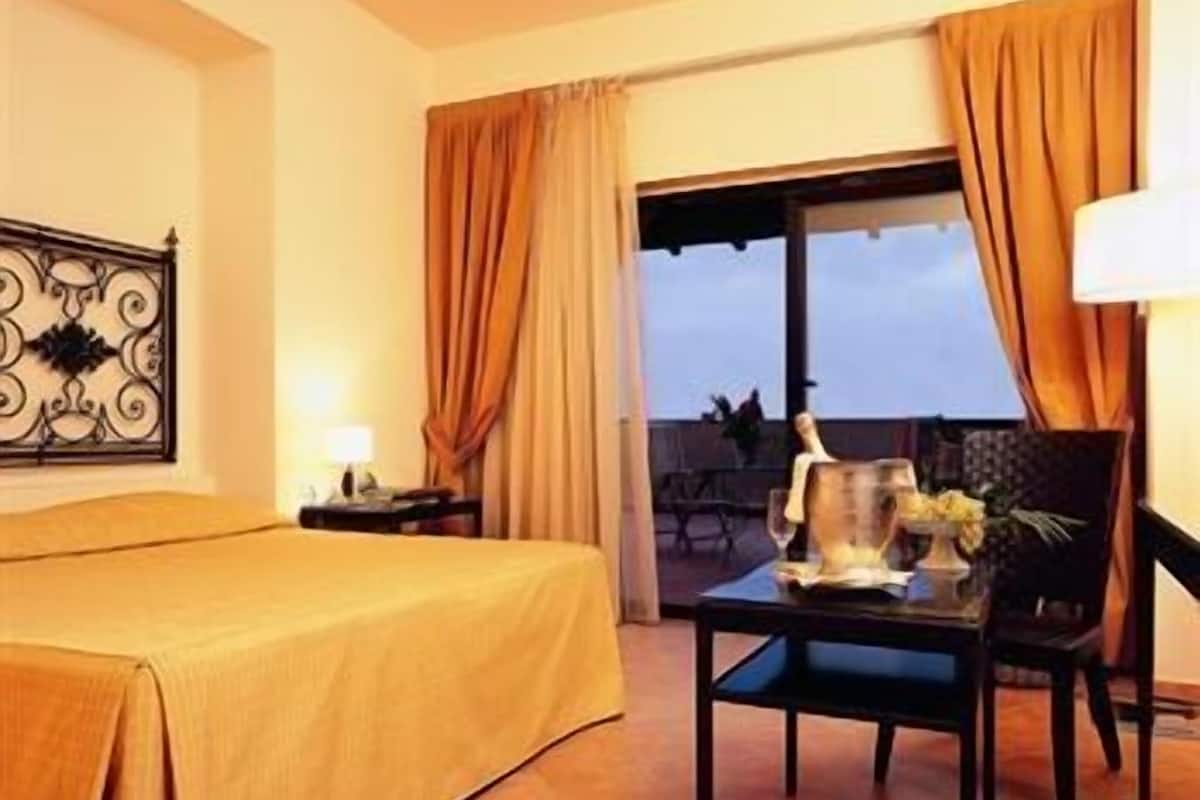 Ein Hotelzimmer mit einem Bett, einem Tisch mit Lampe und Blick durch ein mit Vorhängen drapiertes Fenster.