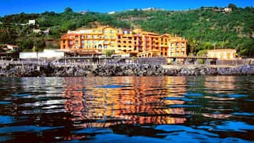 A waterfront hotel with multiple balconies, a stone pier, and a hillside backdrop.