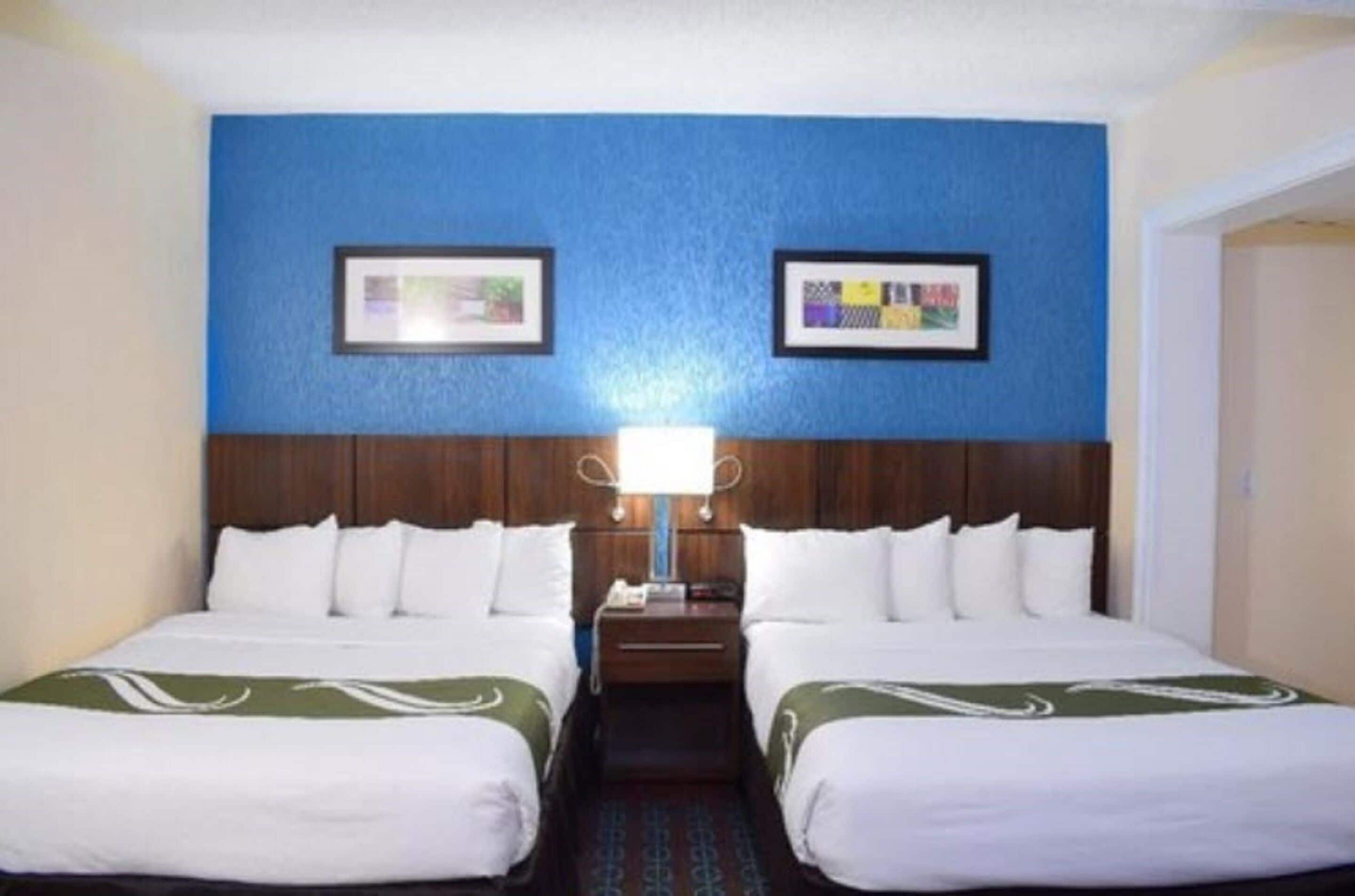 A hotel room with two beds, a wooden headboard, a nightstand, and a wall-mounted lamp.