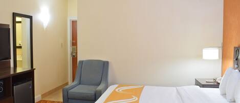 A hotel room with a bed, a chair, a desk with a television, and a nightstand with a lamp.