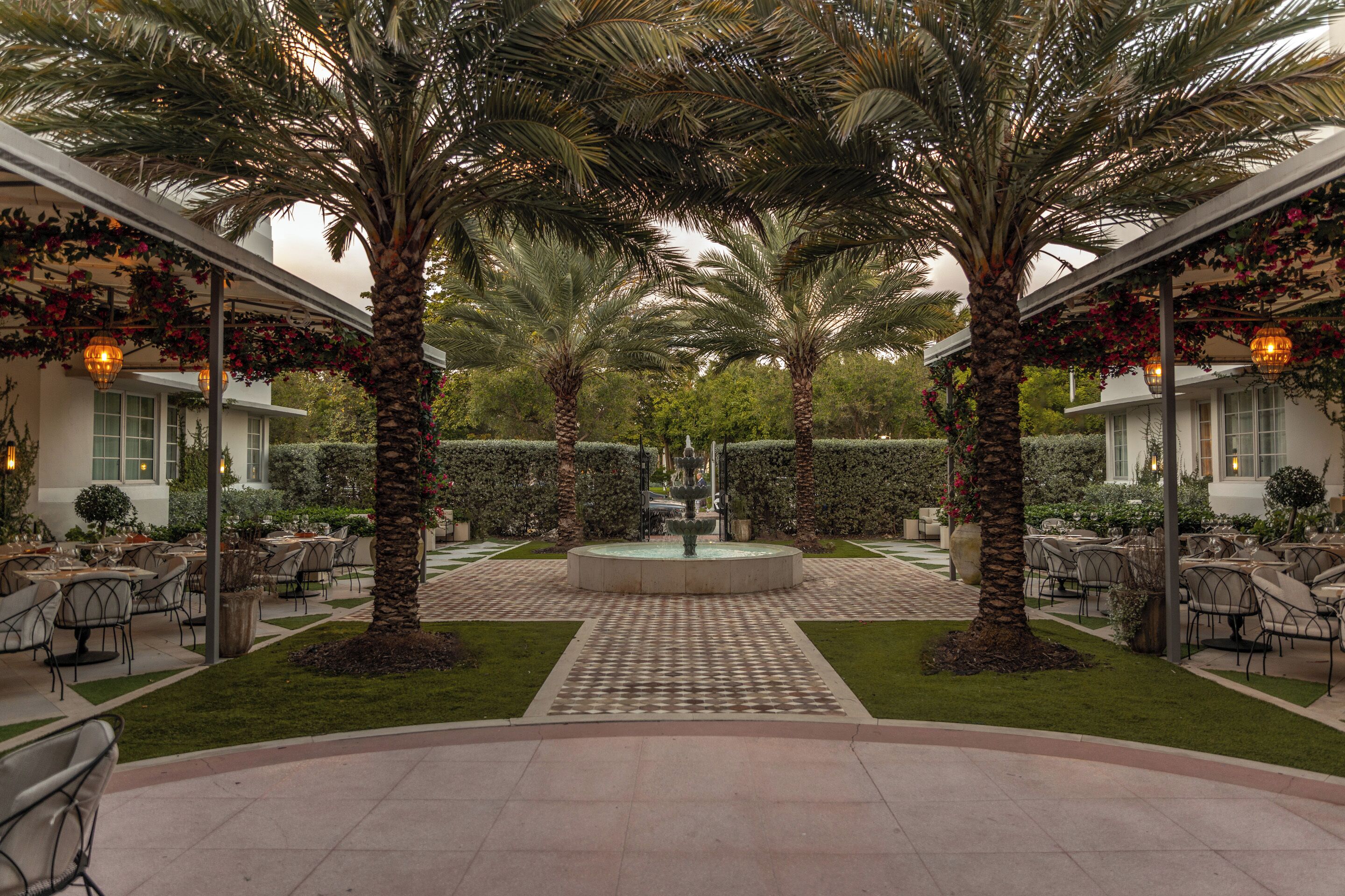 An outdoor dining area with palm trees, a central fountain, and tables and chairs arranged for guests.