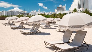 Beach chairs with umbrellas on a sandy beach, with a city skyline in the background. - Liberty Park Hotel South Beach (Miami Beach)