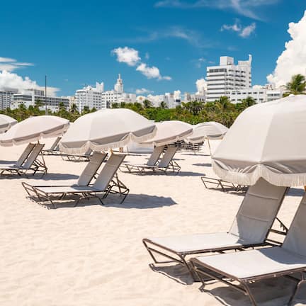 Beach chairs with umbrellas on a sandy beach, with a city skyline in the background.