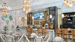 A restaurant interior with wicker furniture, a bar area, and a television displaying a kitchen scene. - Liberty Park Hotel South Beach (Miami Beach)