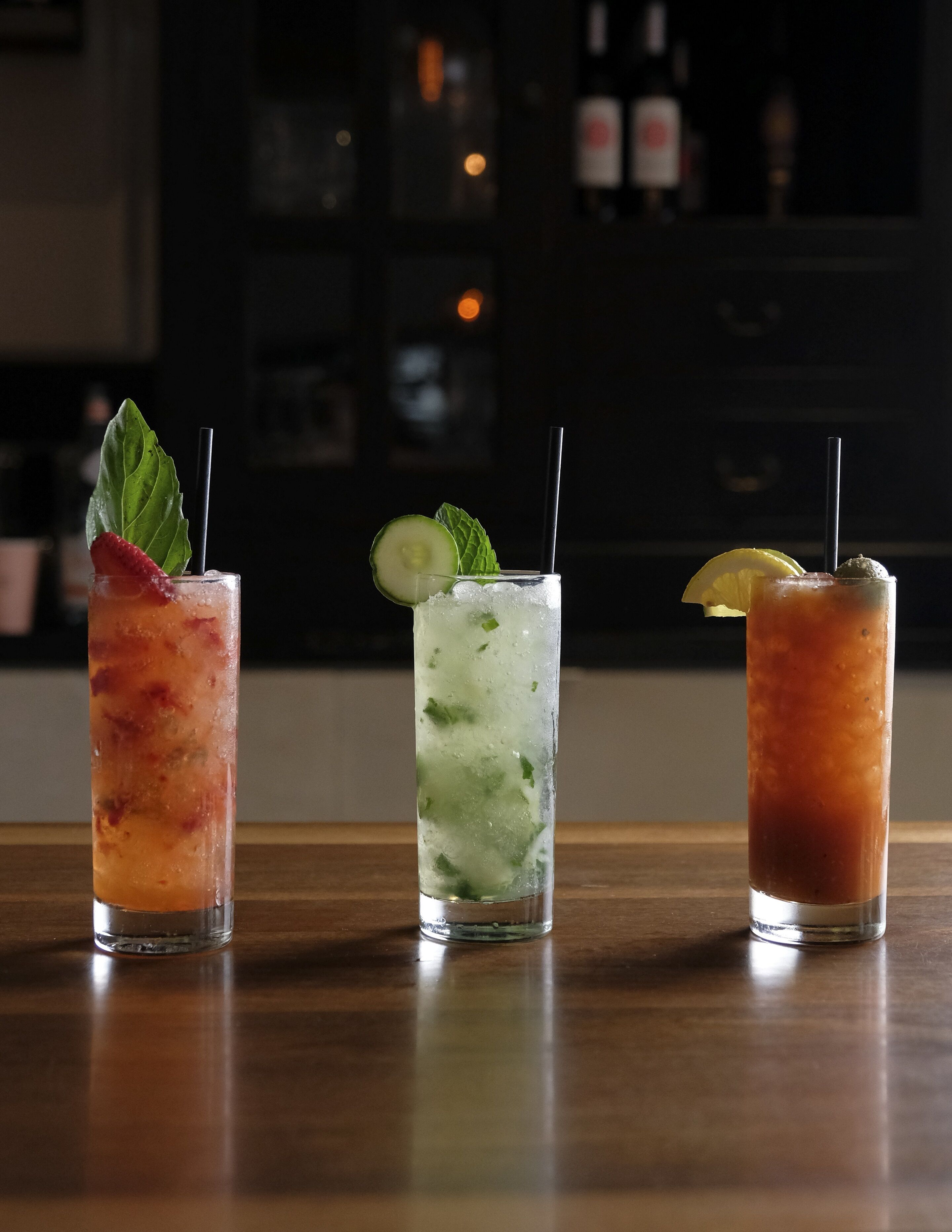 Three tall glasses of iced drinks on a wooden bar counter.